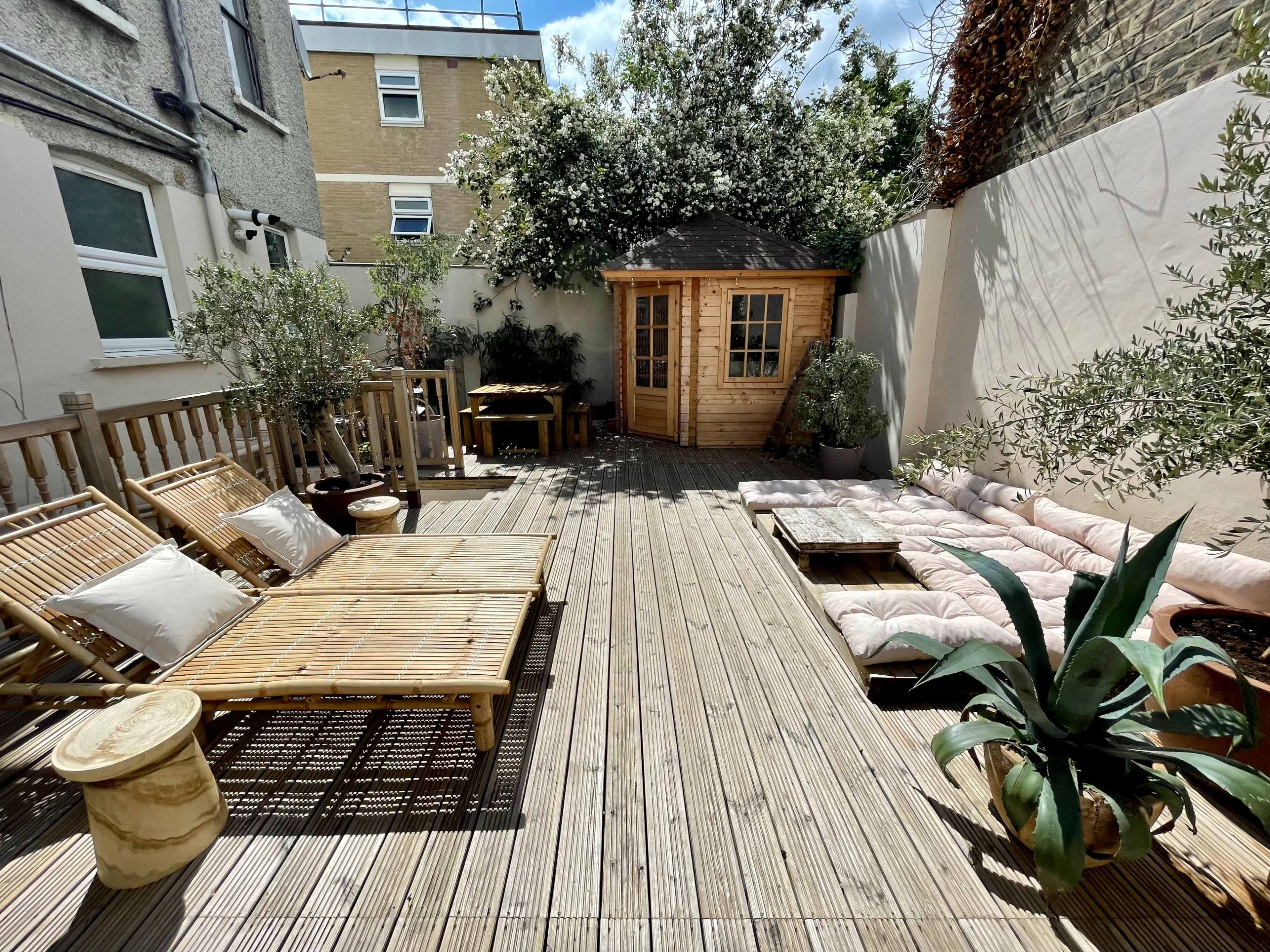 A wooden deck area with lounge seating, a small shed, and potted plants, surrounded by walls and buildings.