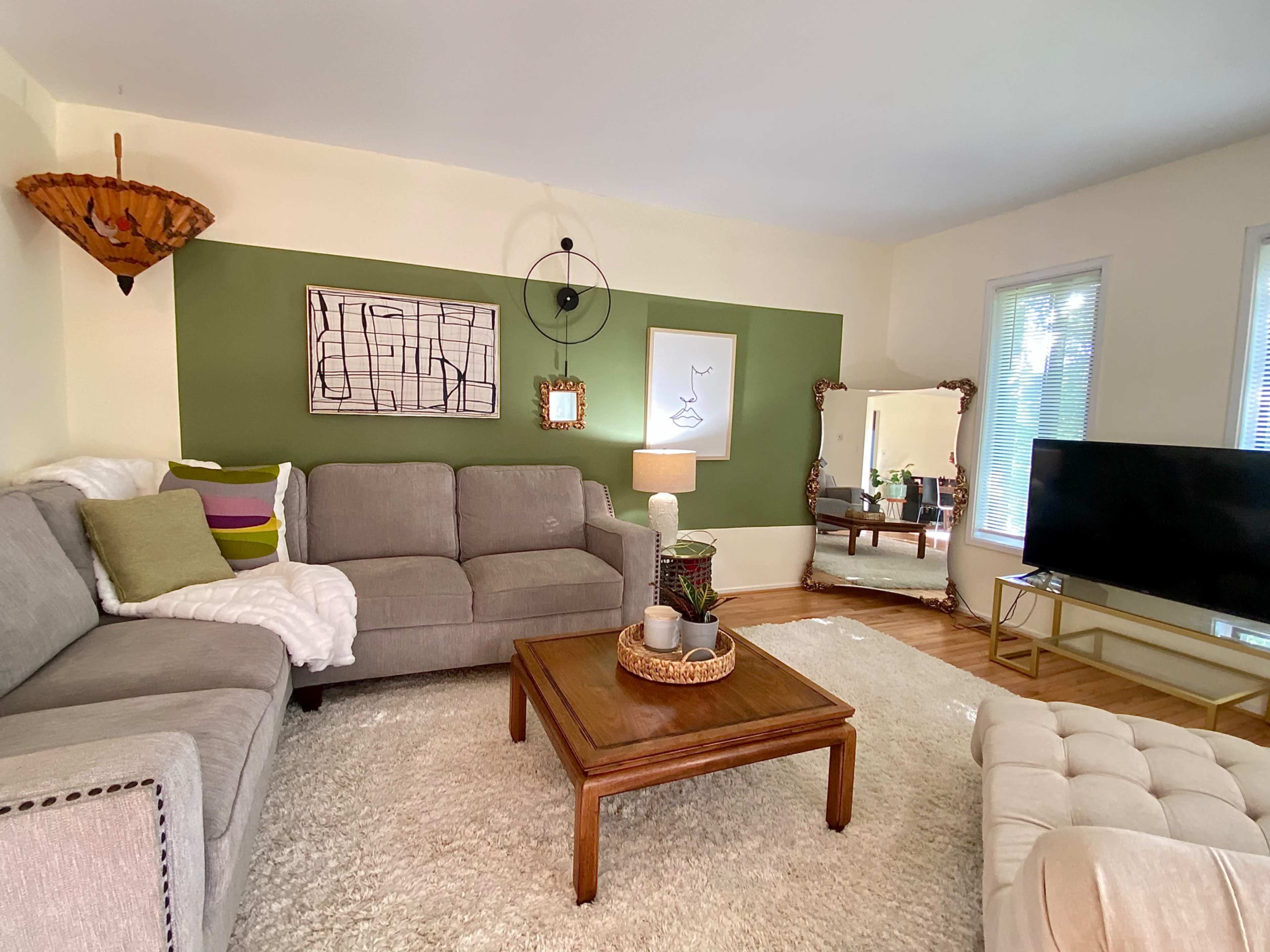A living room featuring a gray sectional sofa, a wooden coffee table, a large mirror, and a television against a wall with a green accent.
