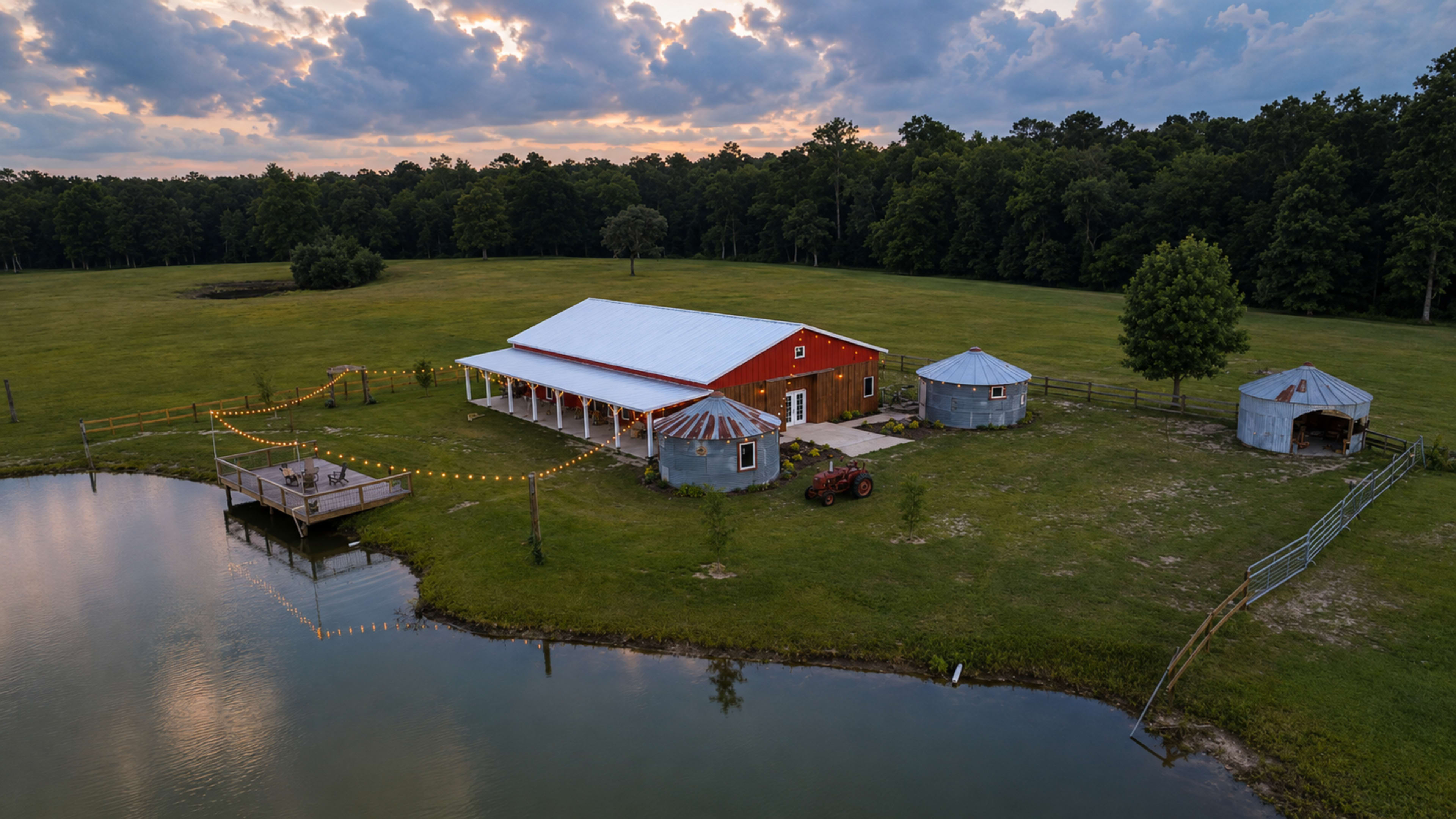 A red barn and outbuildings are situated near a pond in a grassy field, surrounded by trees under a cloudy sky.