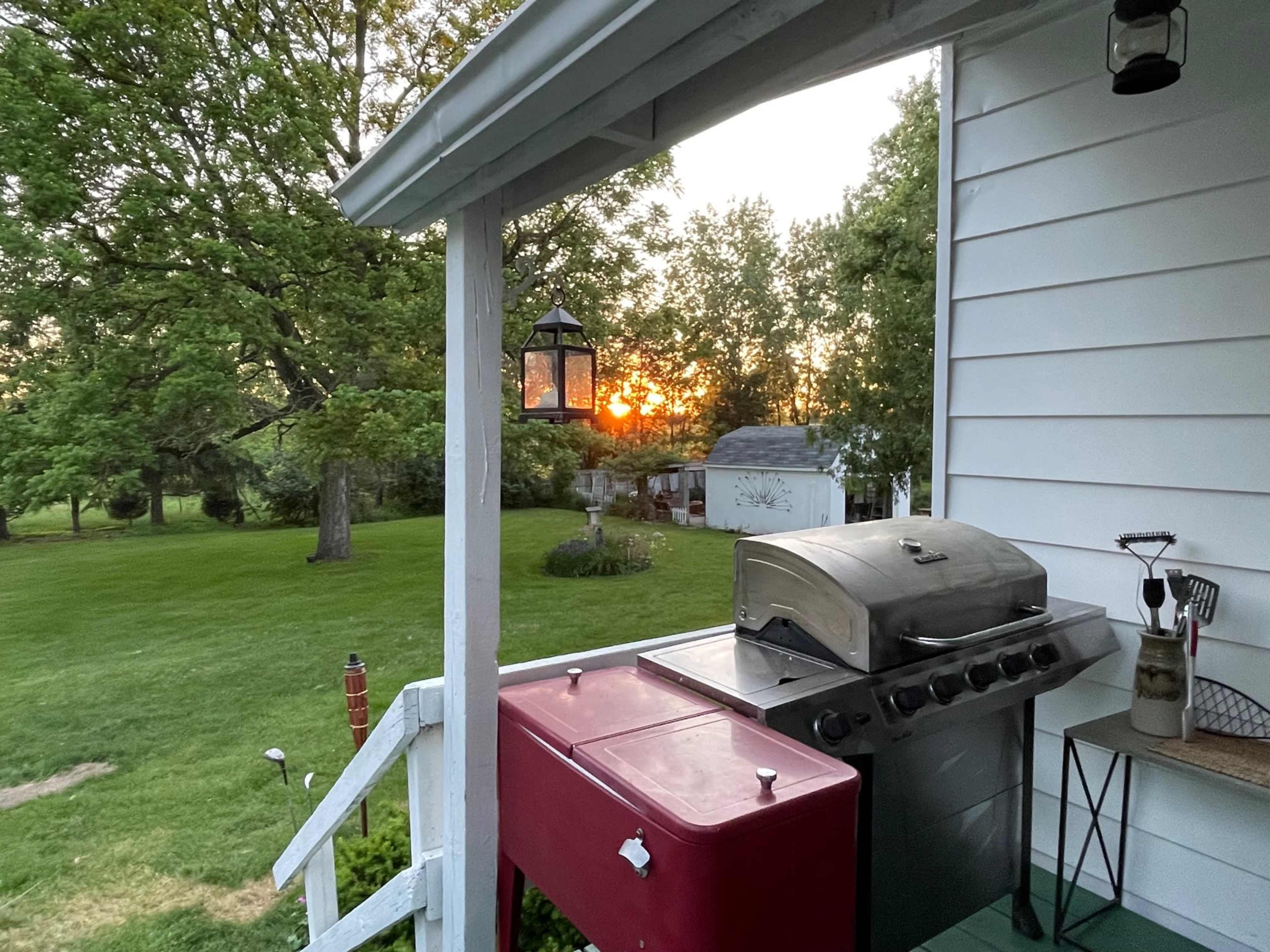 A barbecue grill and storage unit are positioned on a porch overlooking a green lawn with a sunset in the background.