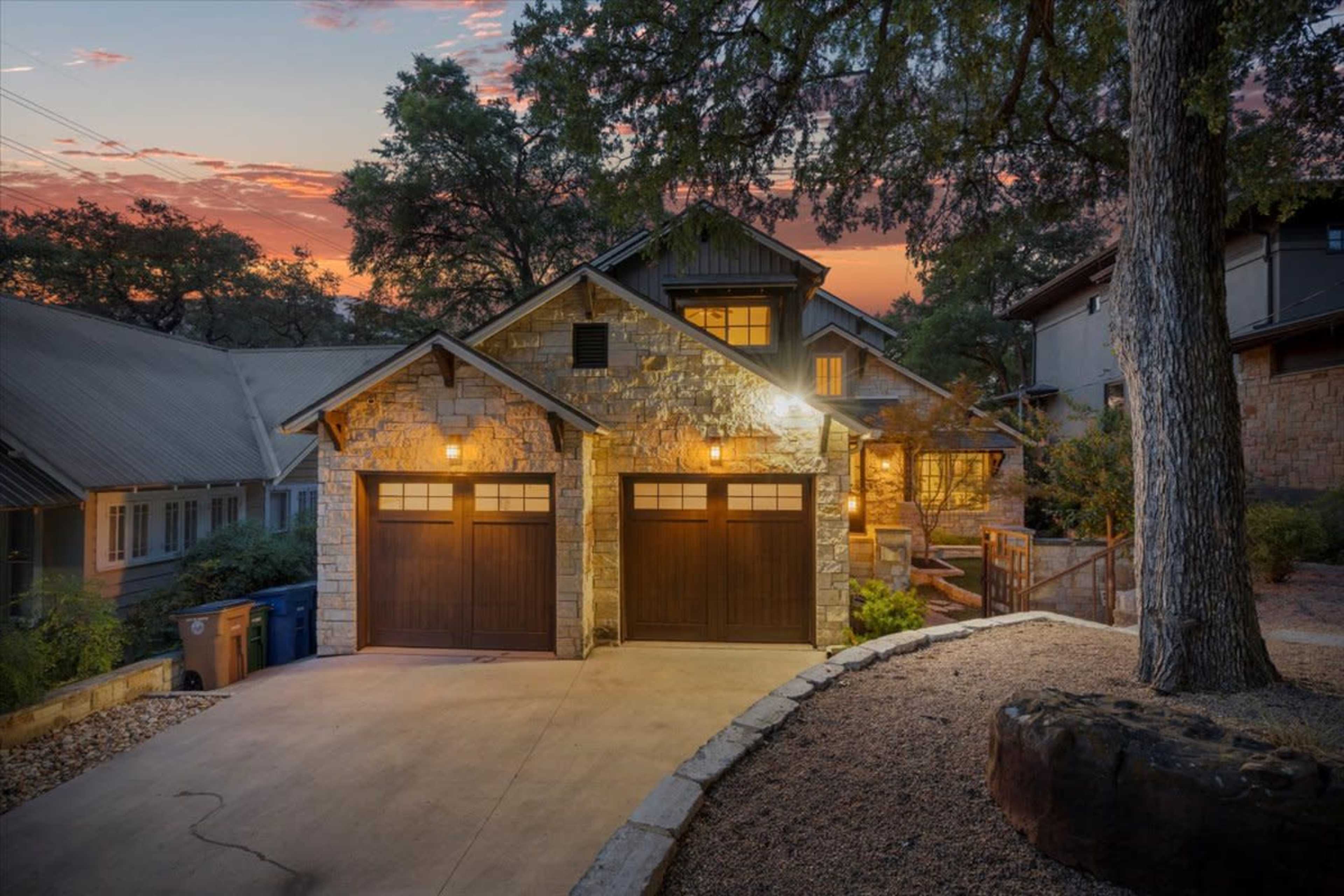 A two-story stone house with a double garage and warm lighting sits amid trees at dusk.