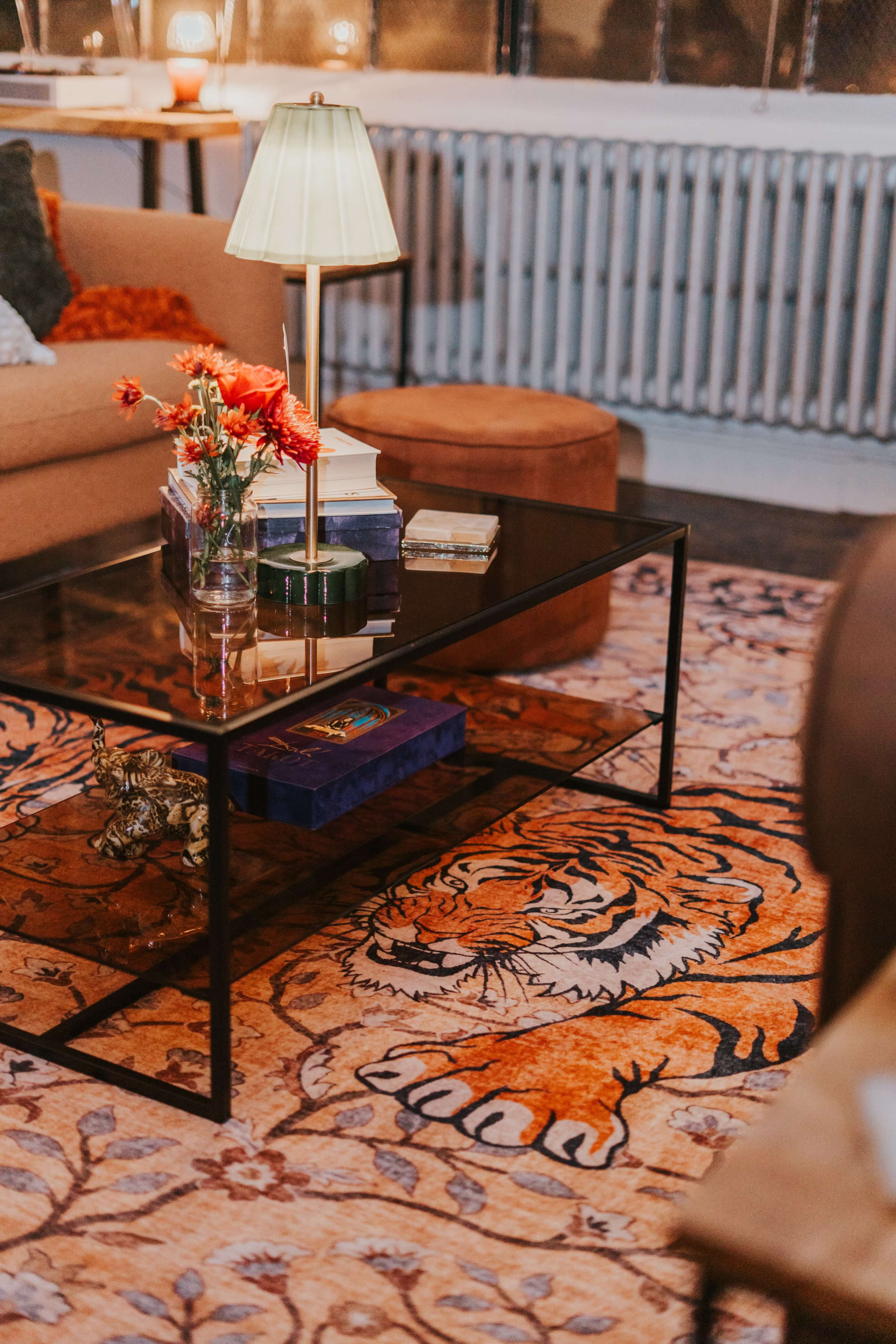 A glass coffee table with flowers sits on a colorful rug featuring a tiger, surrounded by a sofa and lamp in a cozy living room setting.