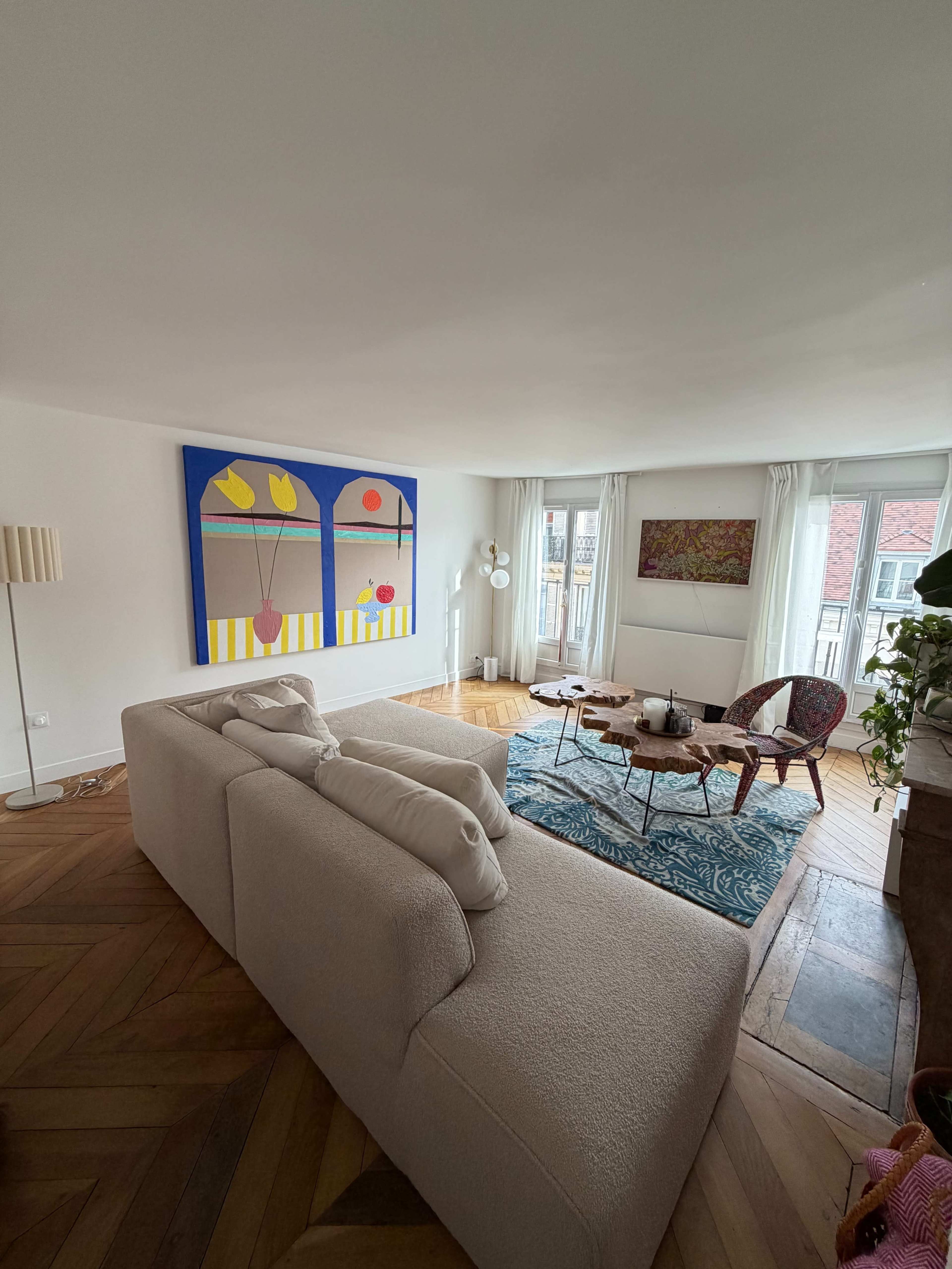The image shows a bright and modern living room with a large abstract painting, a light-colored sofa, a coffee table, and a patterned area rug.