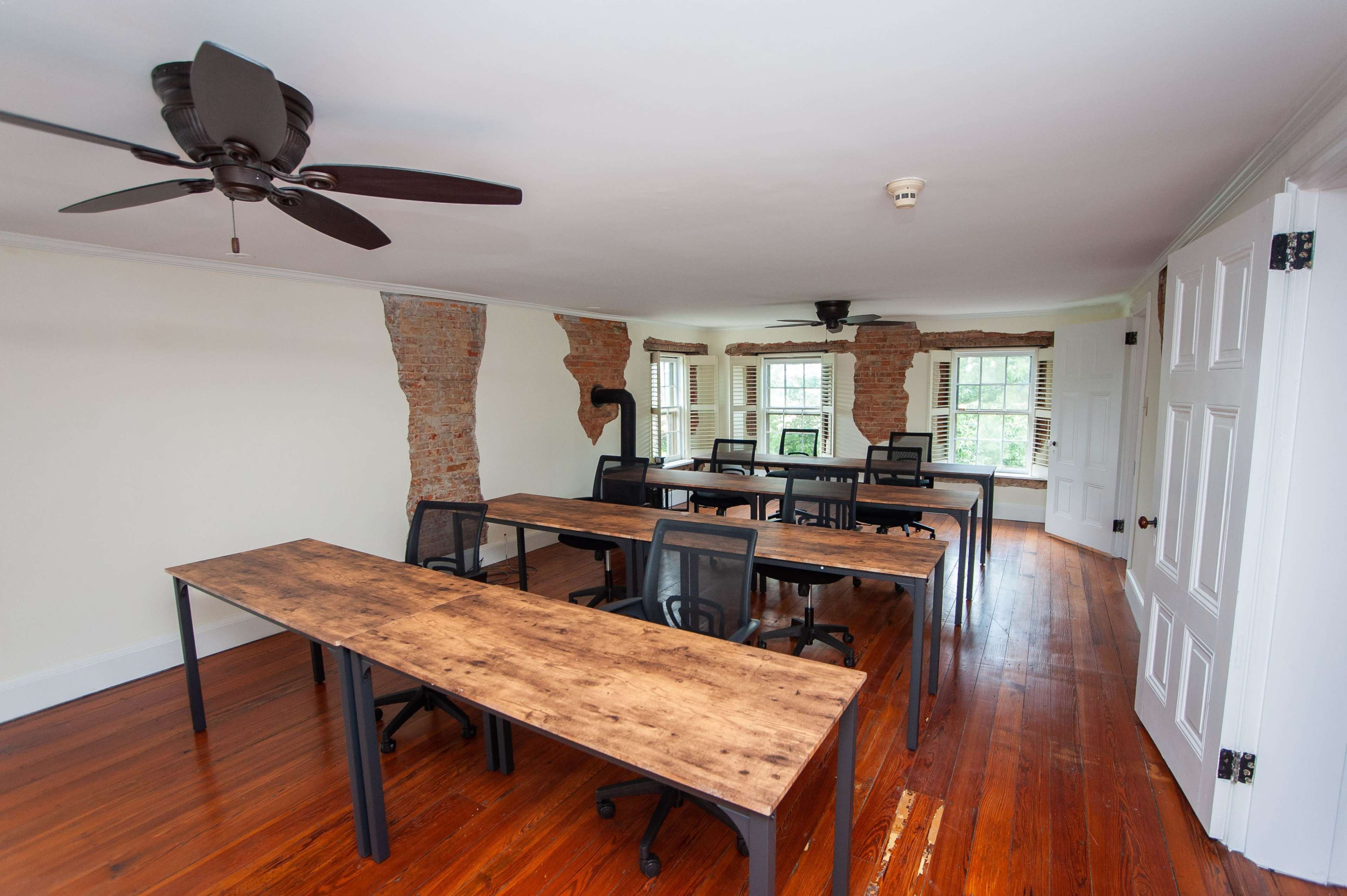 The room features several wooden tables arranged with office chairs, positioned under a ceiling fan, and includes exposed brick walls.