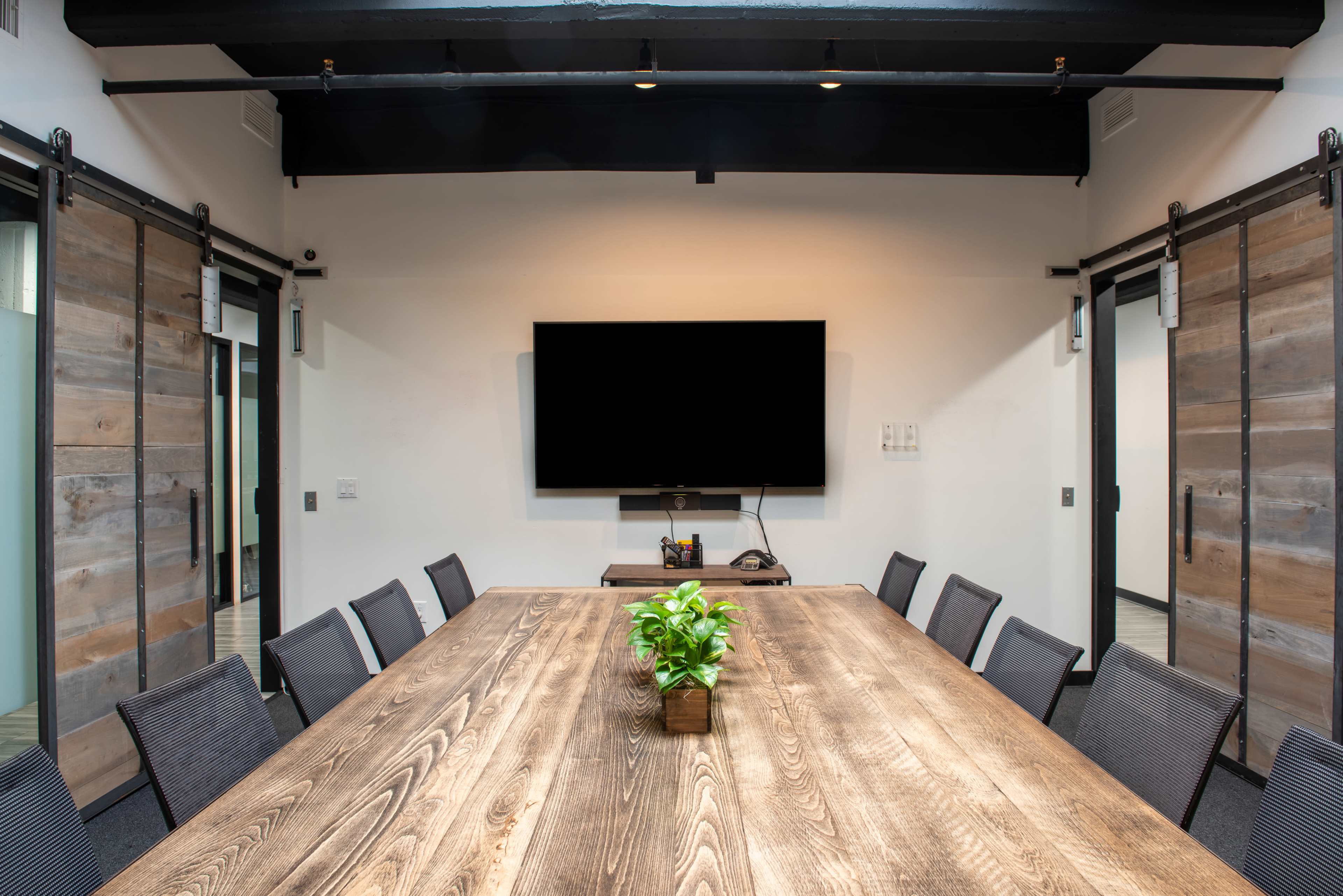 A modern conference room features a large wooden table surrounded by chairs, with a wall-mounted television and sliding barn doors.