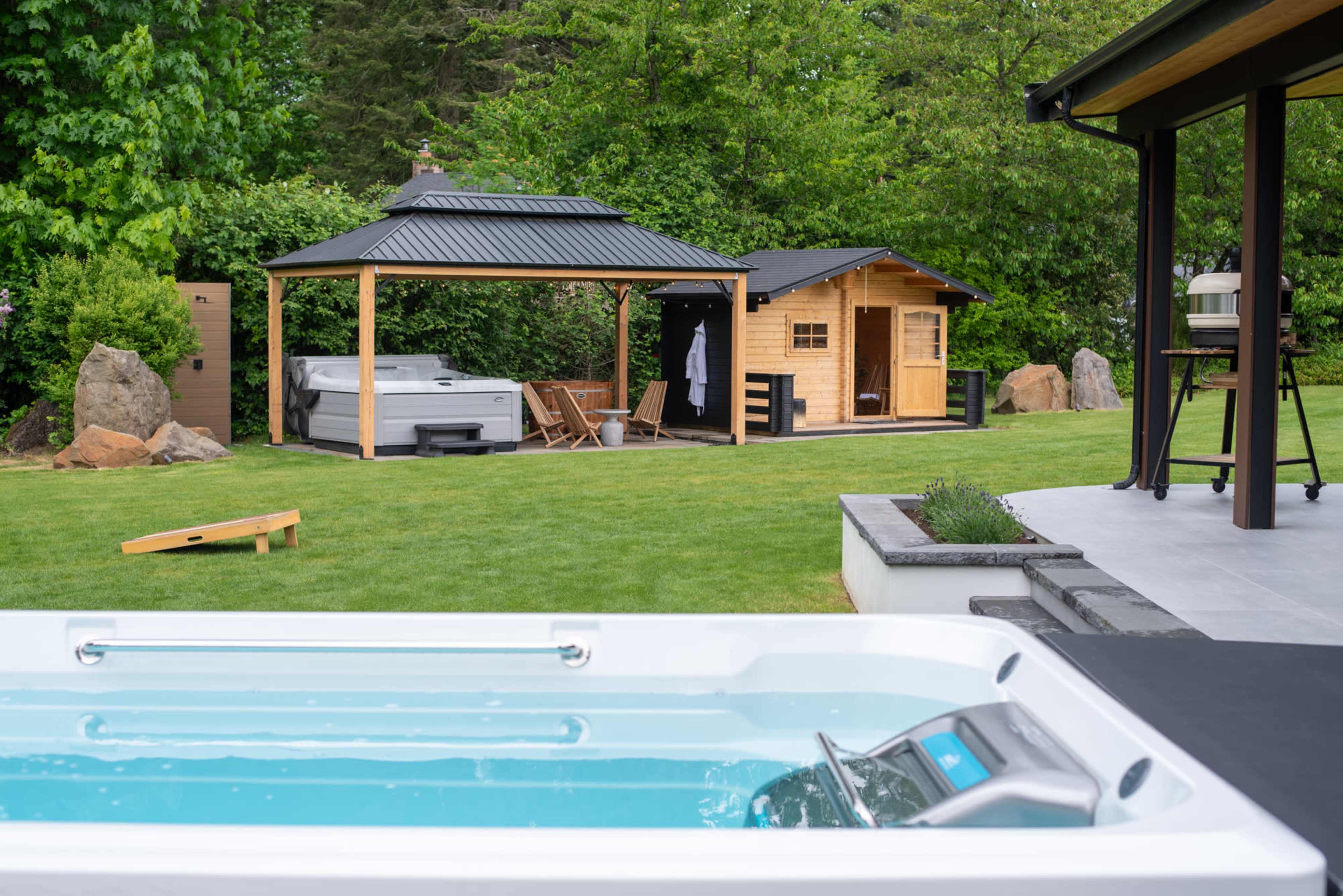 The scene features a backyard with a hot tub, a wooden sauna, and a small shed, all surrounded by lush green grass and trees.