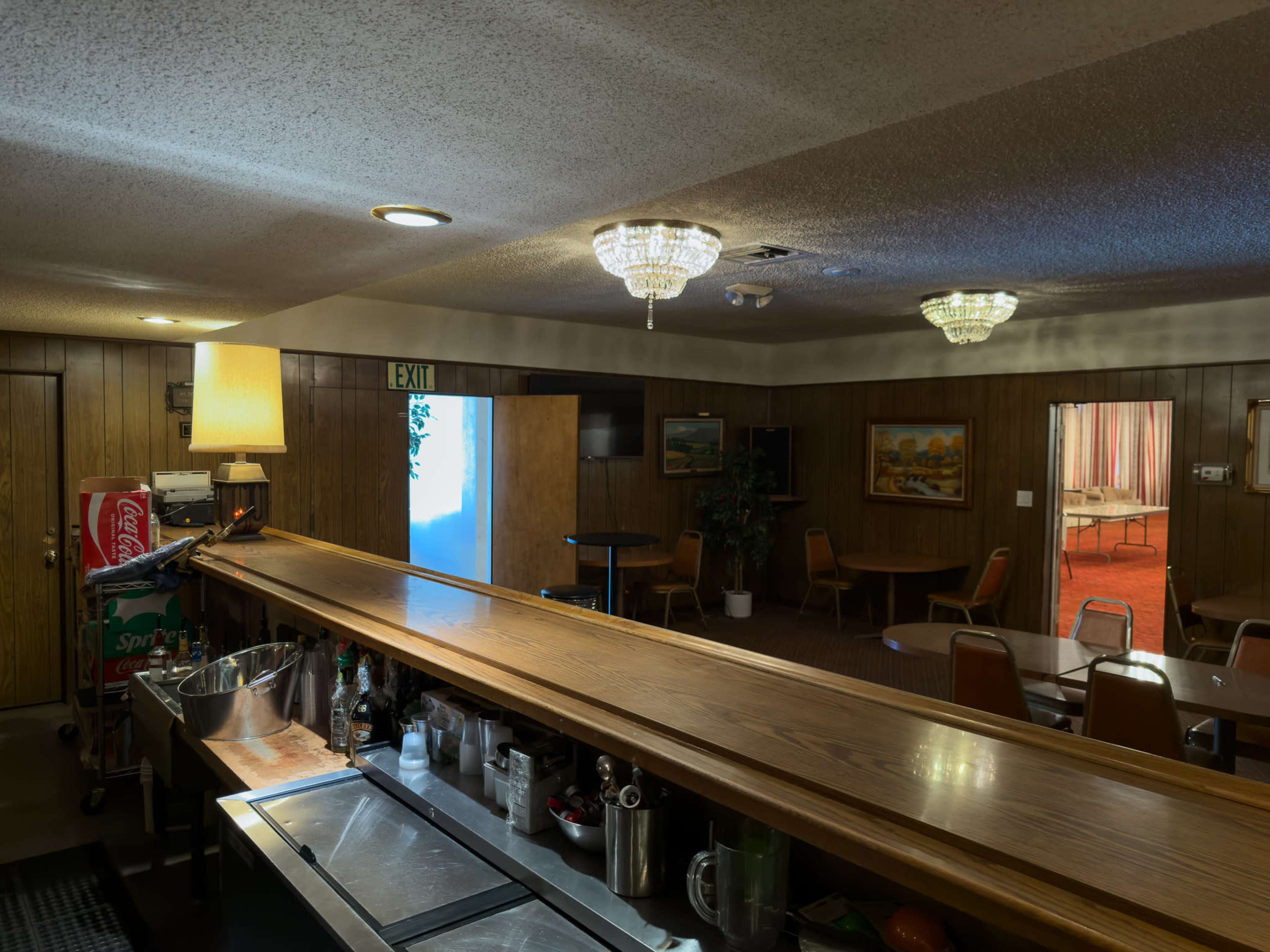 The image shows a dimly lit bar area with a wooden counter, surrounded by brown paneling and featuring tables and chairs in a separate room.