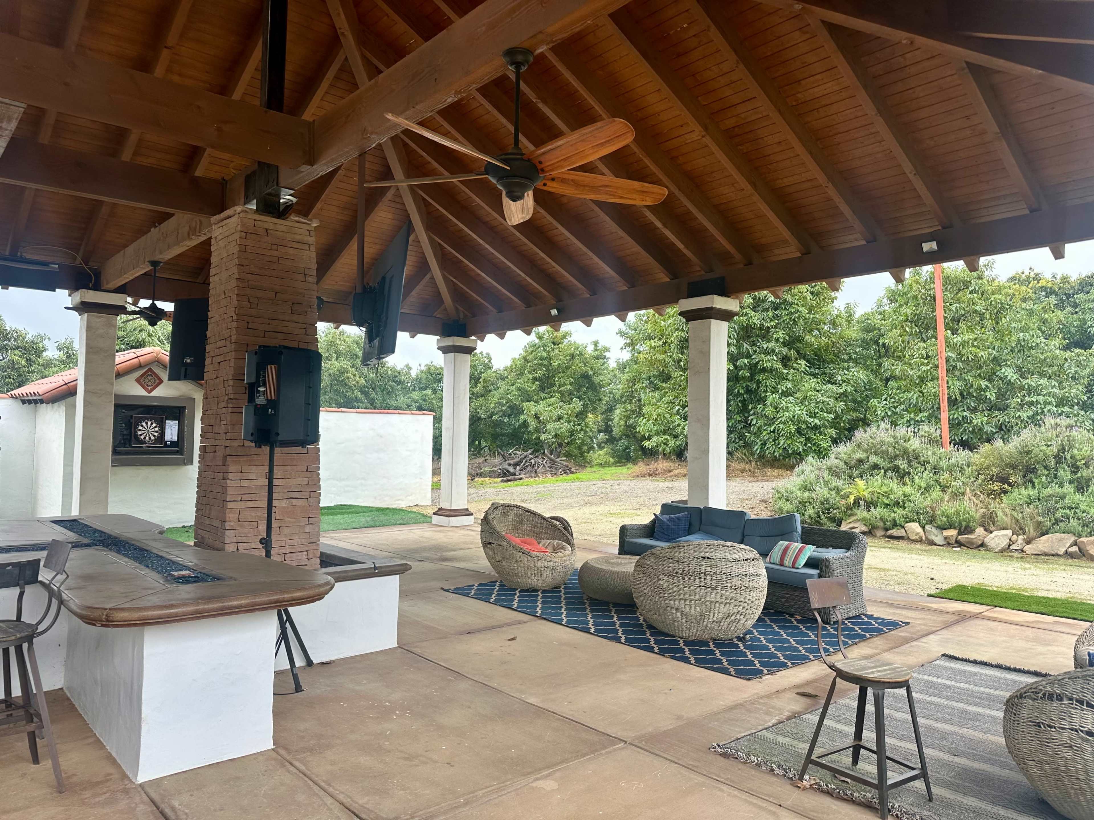 The image shows a spacious outdoor patio area with a wooden ceiling, featuring a seating arrangement with a couch and poufs, along with a bar counter and greenery in the background.