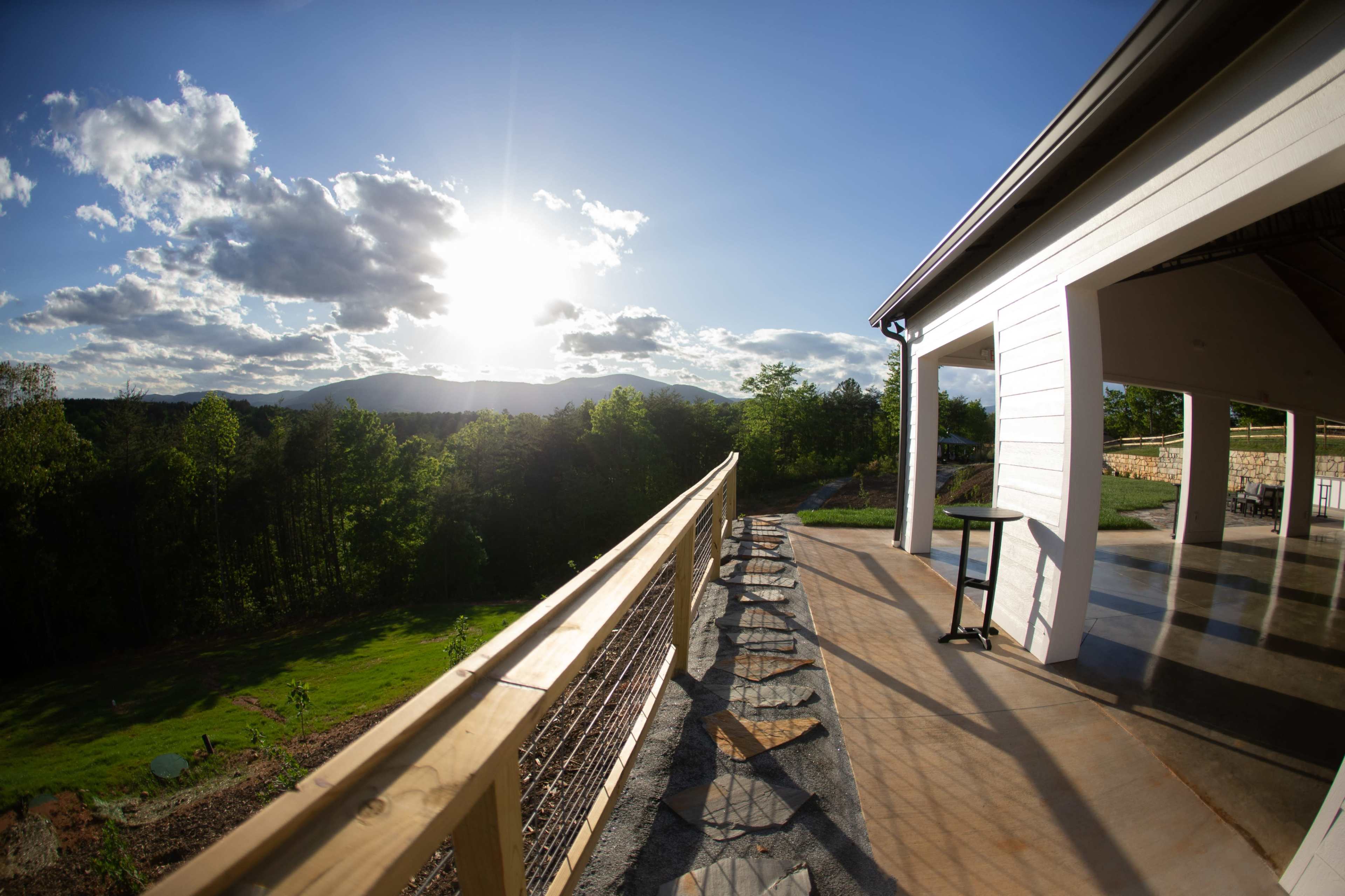 A spacious balcony overlooks a green landscape and distant mountains under a bright sky filled with clouds.