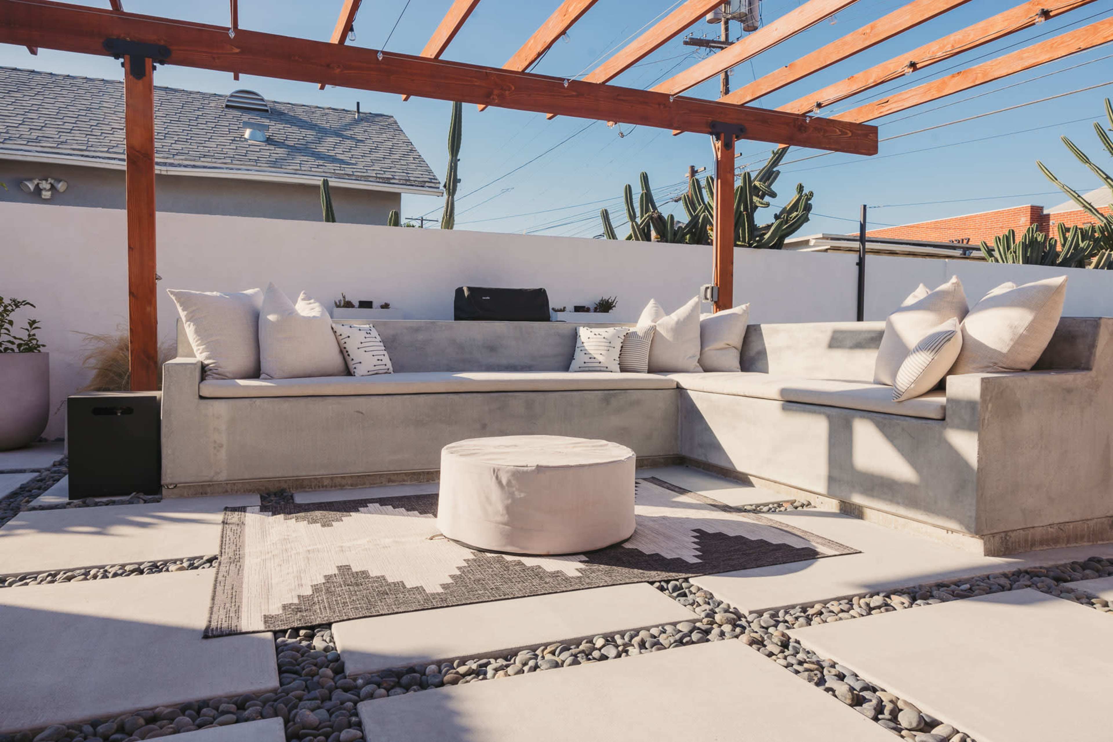 A modern outdoor seating area with a concrete sofa, decorative pillows, a circular table, and a gravel patio under a wooden pergola.