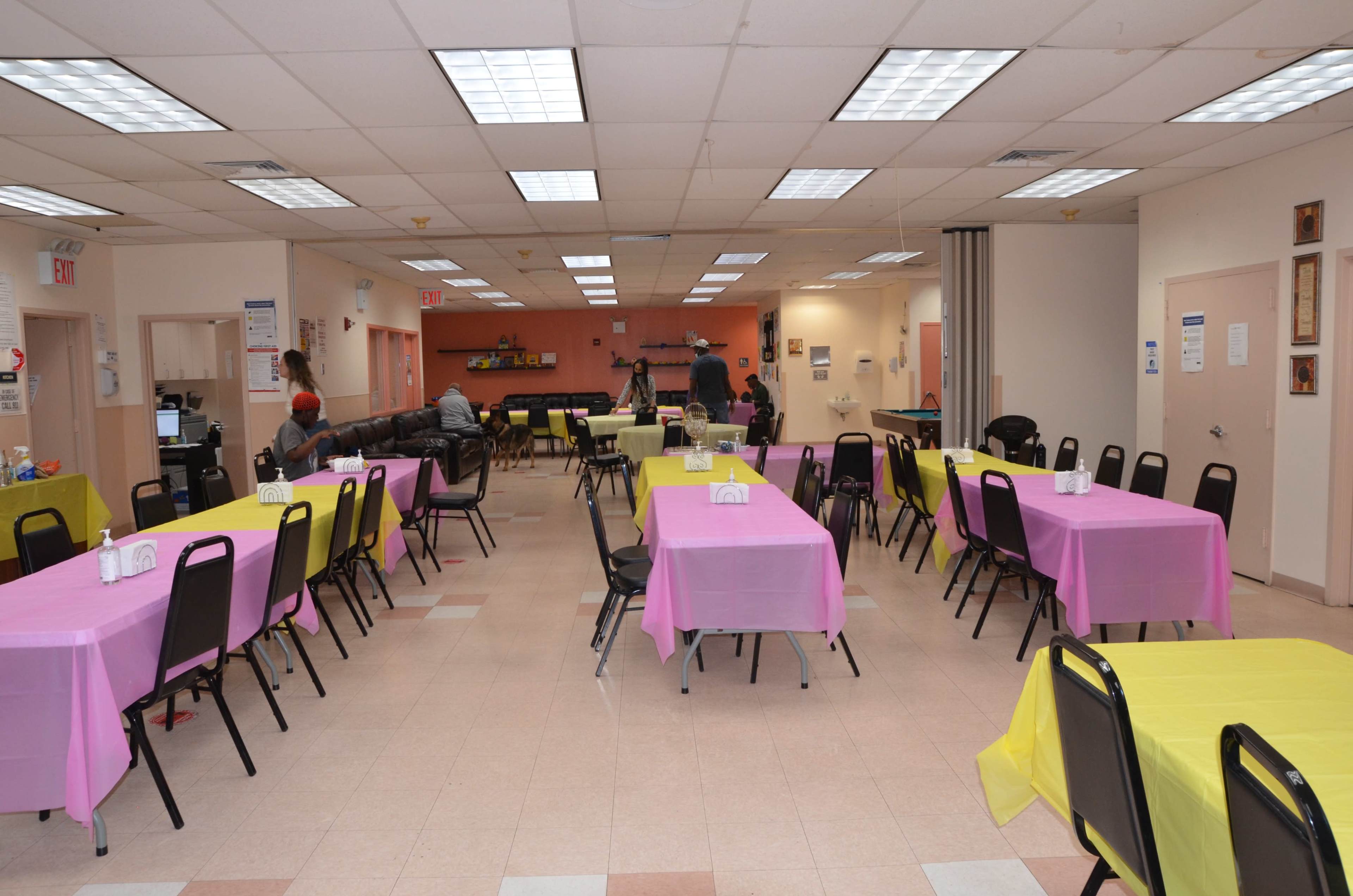 A spacious community room features several tables covered with pink and yellow tablecloths, arranged for an event.