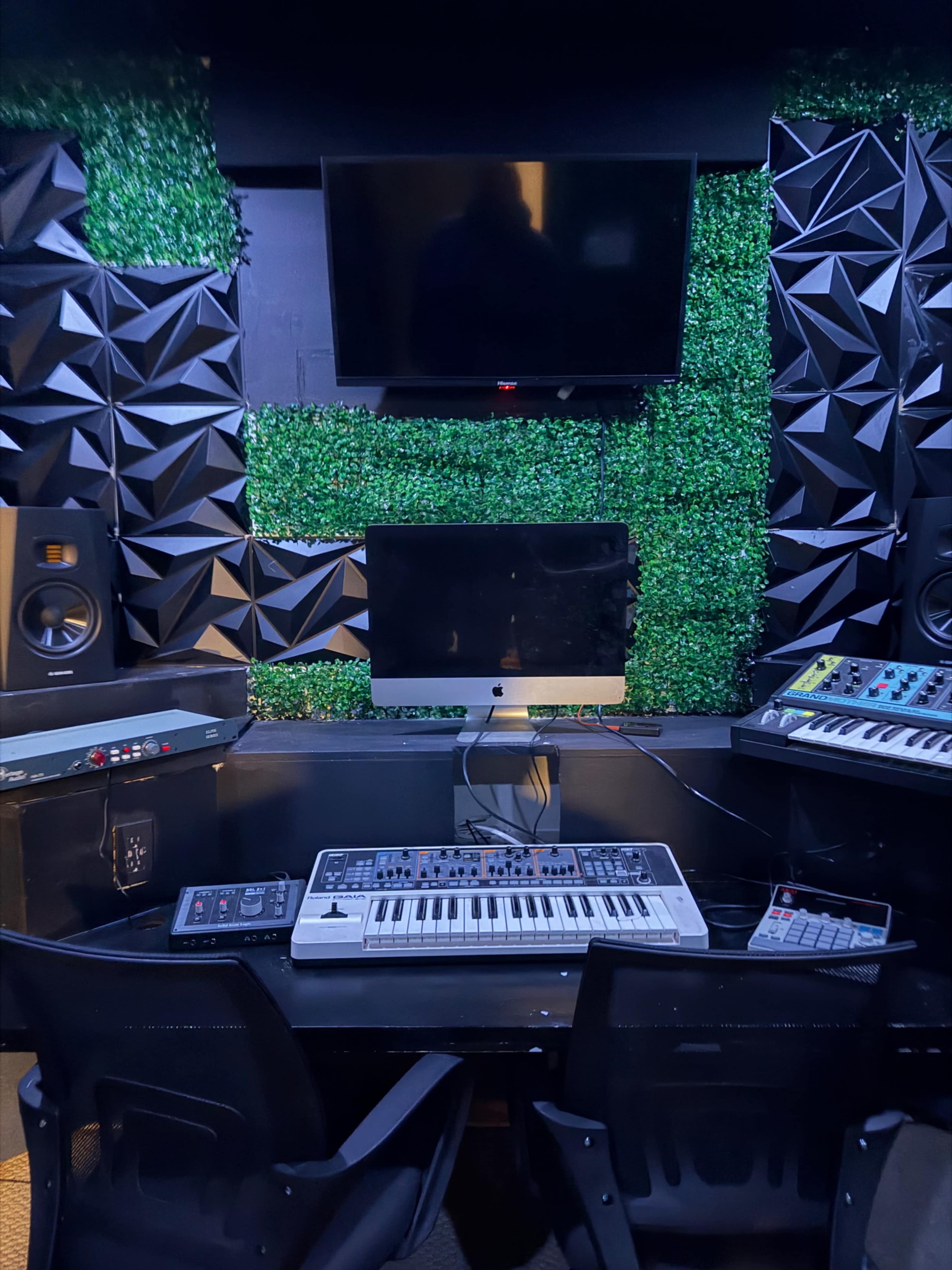 The image shows a music production studio featuring a keyboard, a computer display, and sound equipment, surrounded by soundproofing panels and a green wall.