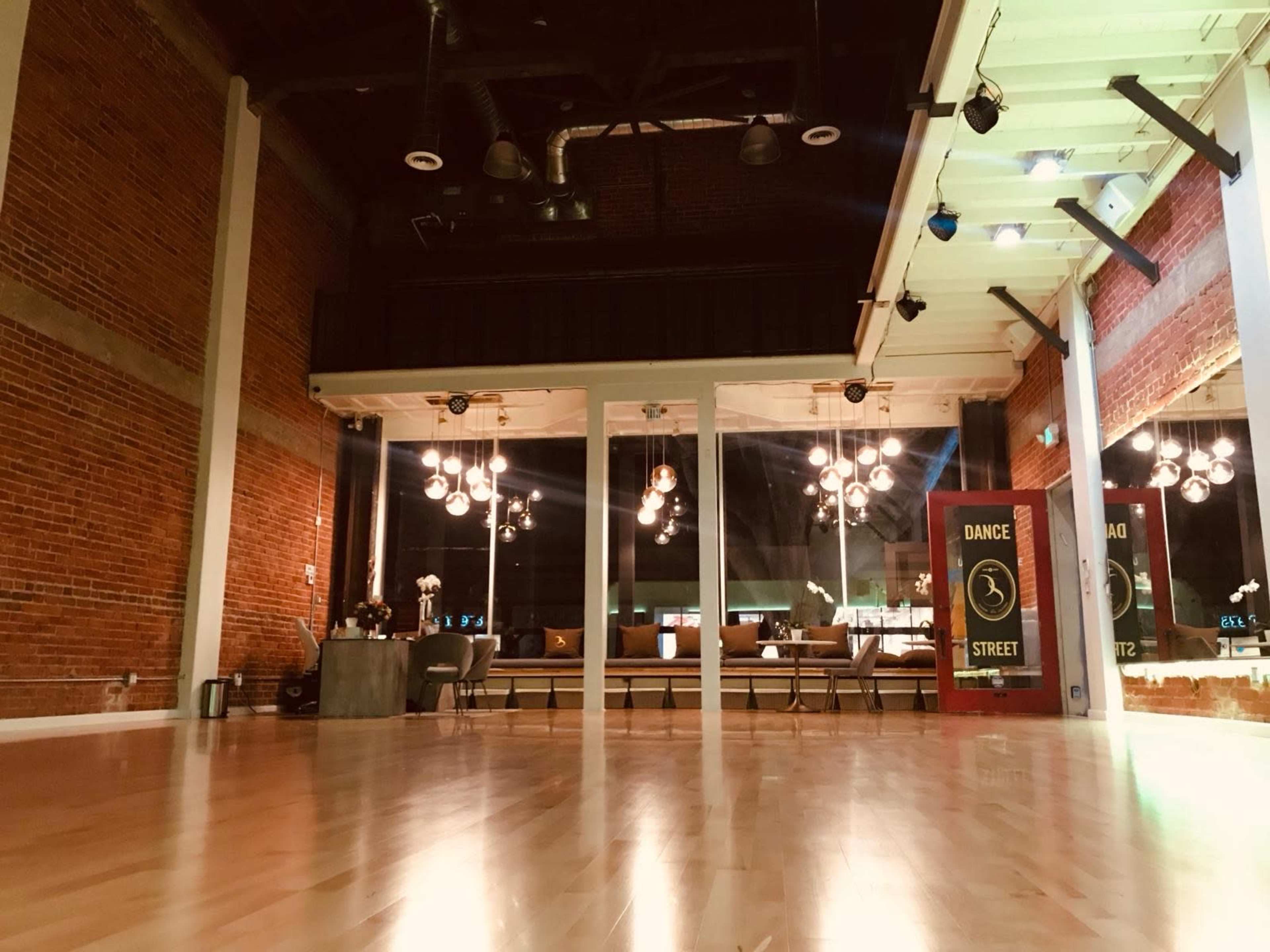The image shows an empty dance studio with exposed brick walls, wooden flooring, and a variety of hanging lights.