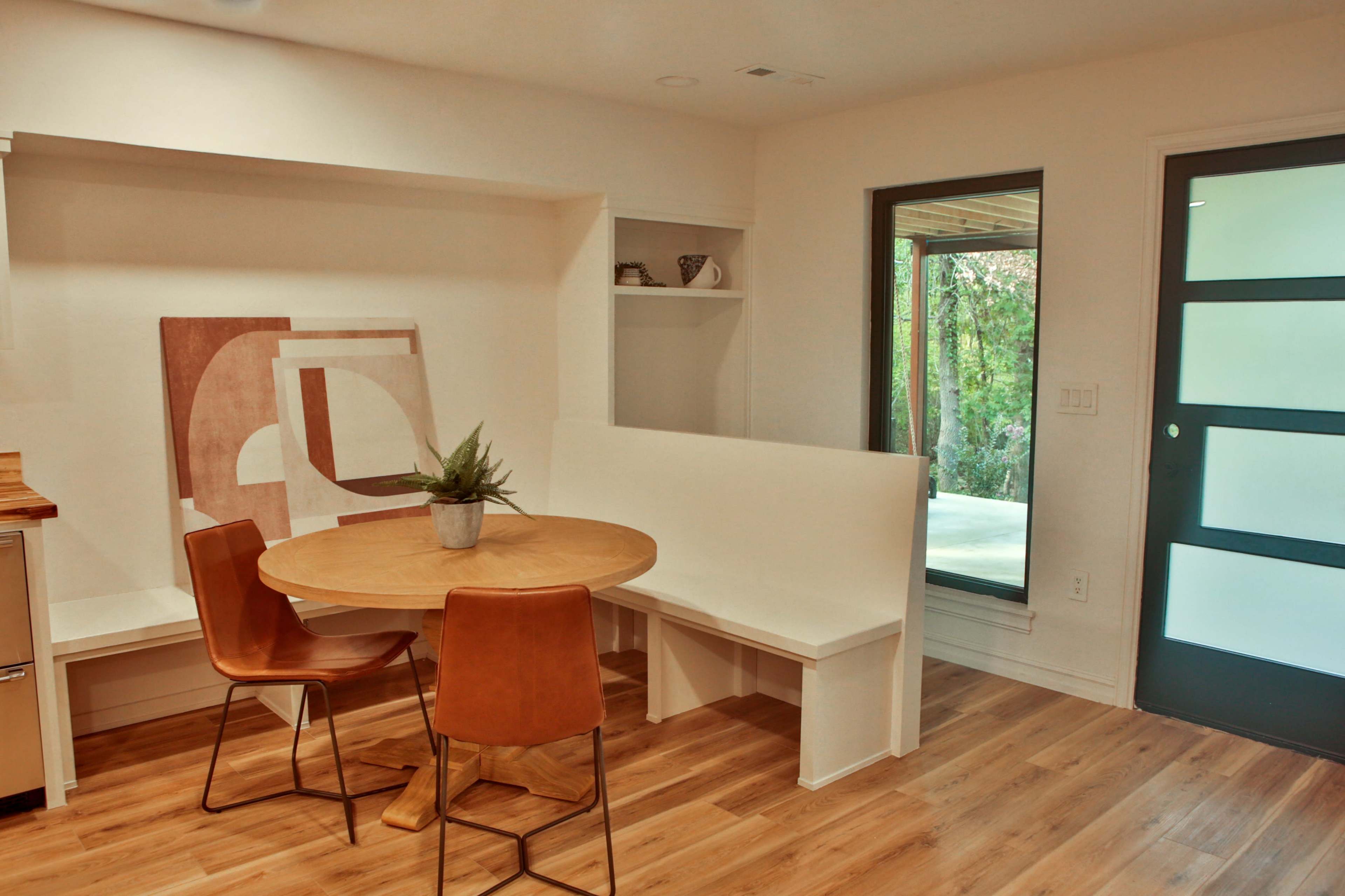 A small dining area features a round wooden table surrounded by two brown chairs and a built-in bench, with a doorway leading to an outdoor space visible through a large window.