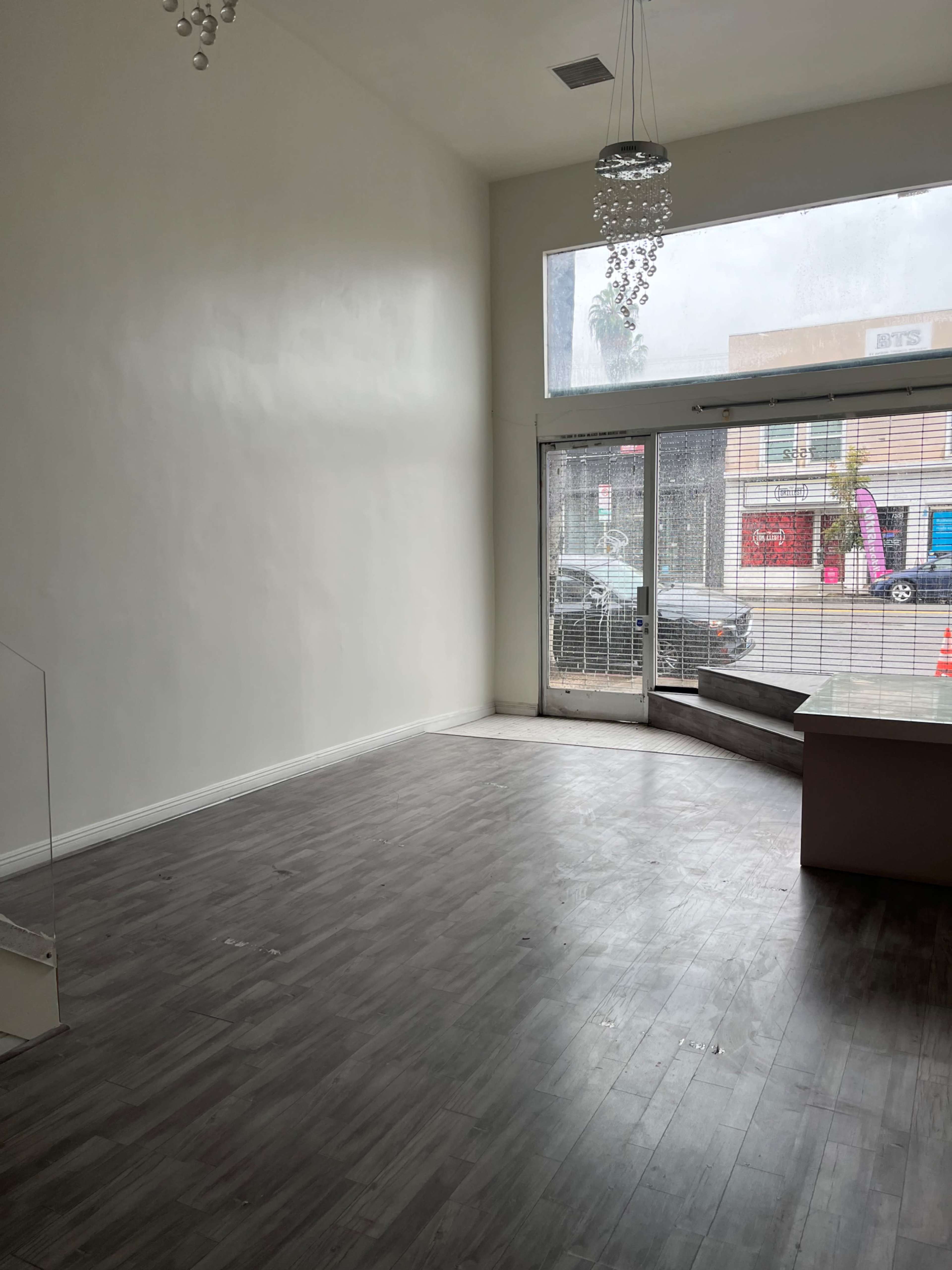 The image shows an empty retail space with a large window and a glass door, featuring a gray floor and minimal furnishings.