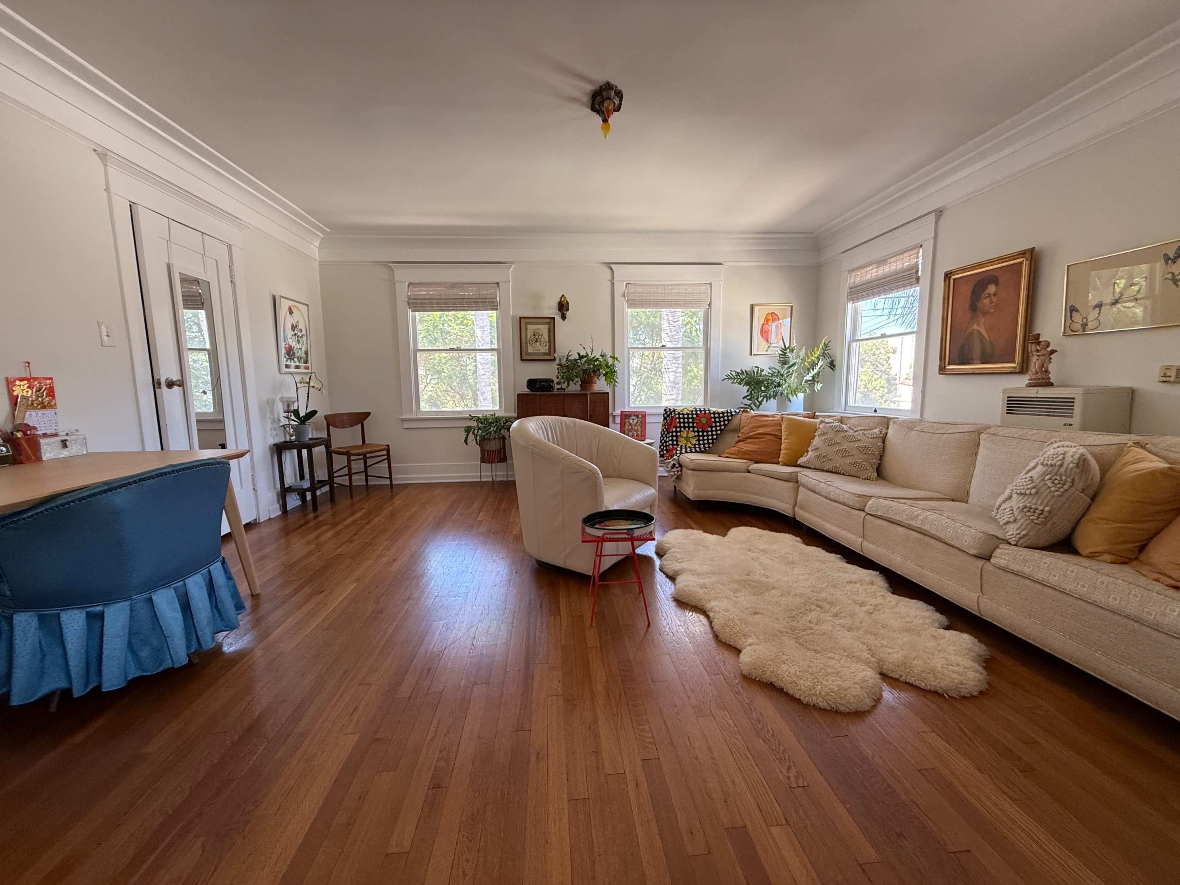 The living room features a large beige sectional sofa, a round black table, and several plants, with large windows allowing natural light to fill the space.