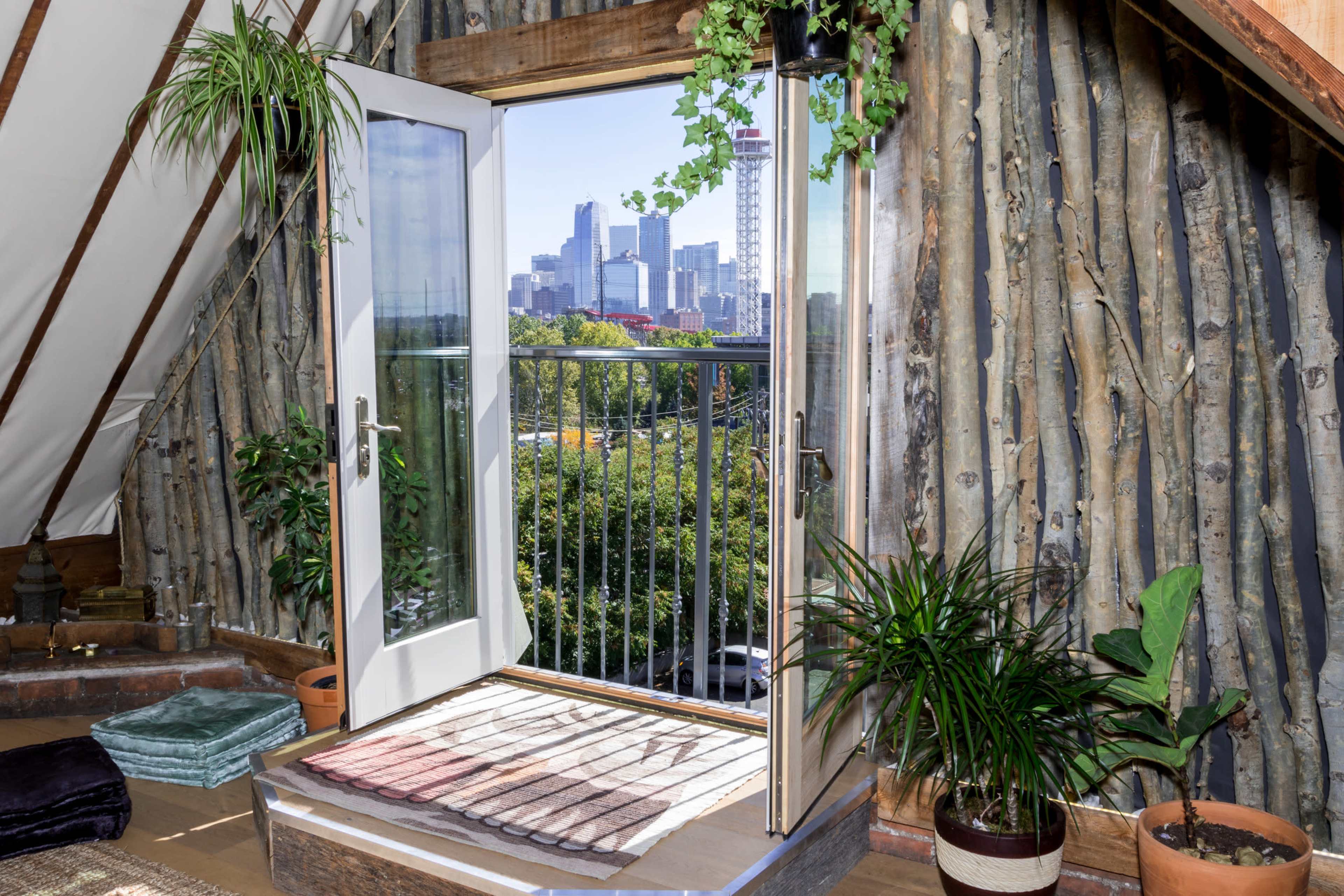A wooden balcony door opens to a view of a city skyline, with greenery and potted plants inside a cozy room.
