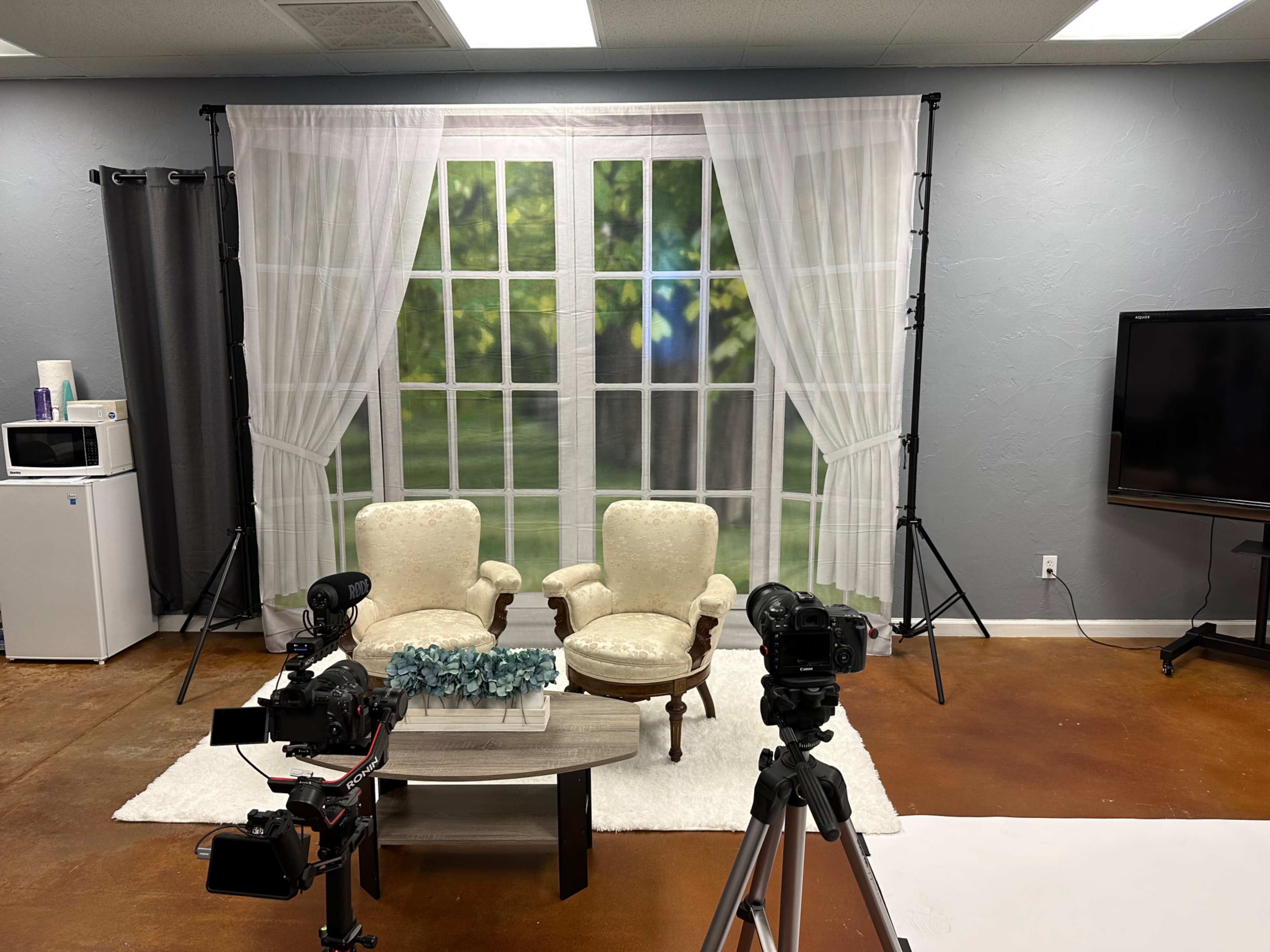 The image shows a filming set with two armchairs facing a coffee table, flanked by cameras, a white rug, and a backdrop of a window.