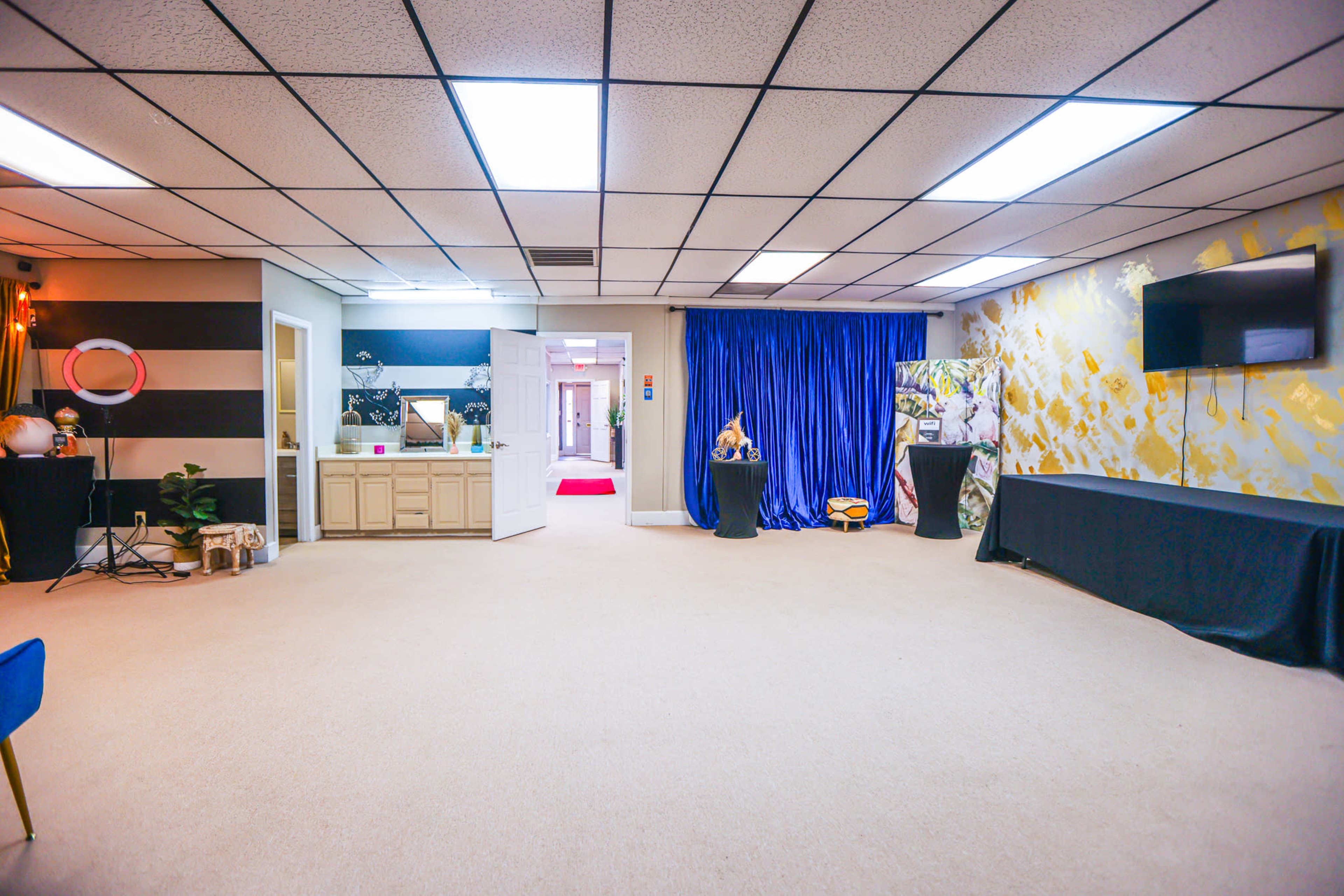 The image shows a spacious room with beige carpet, a visible entrance leading to a red carpet hallway, and blue curtains on one side, along with various furniture and decorations.