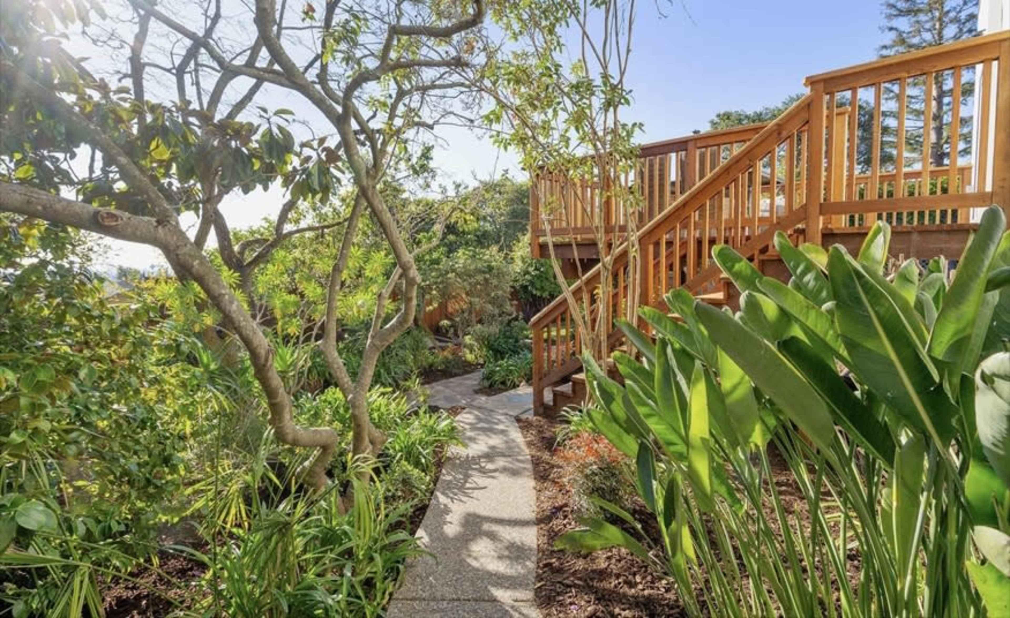 A stone pathway winds through a lush garden, leading up to a wooden staircase surrounded by greenery.