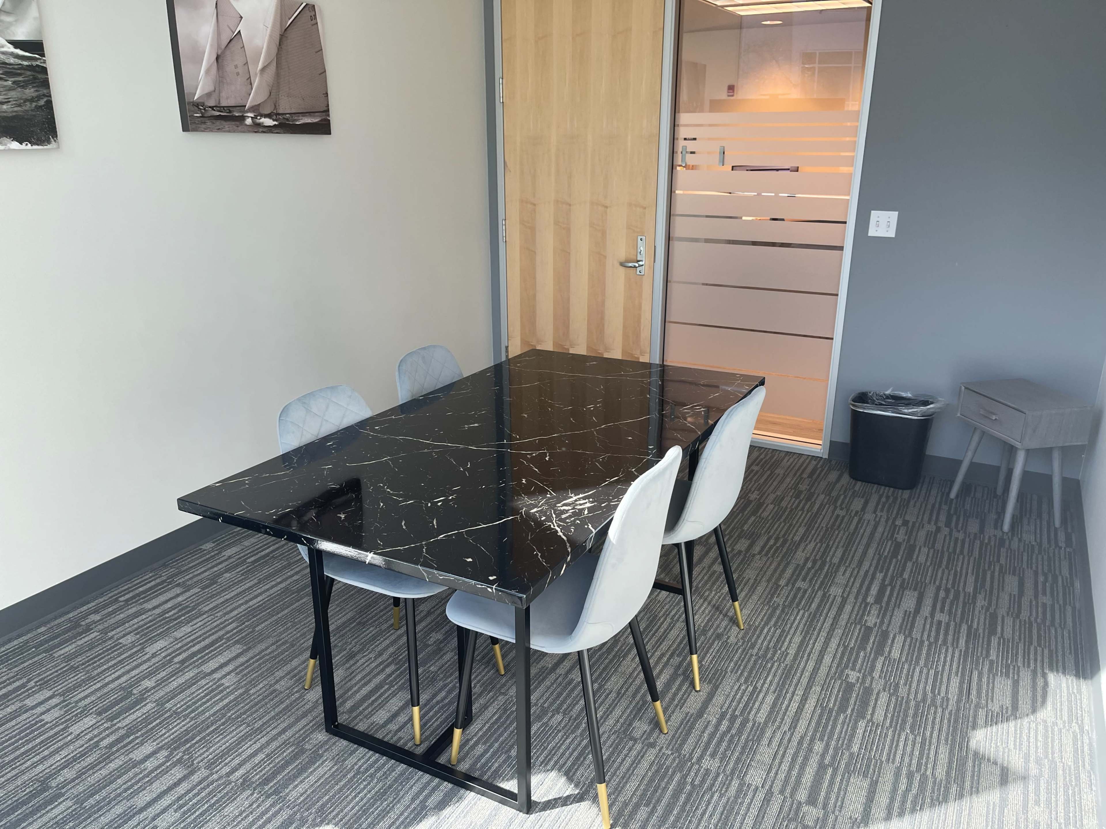 A black marble table with four gray chairs is situated in a well-lit office space next to a glass-paneled door.