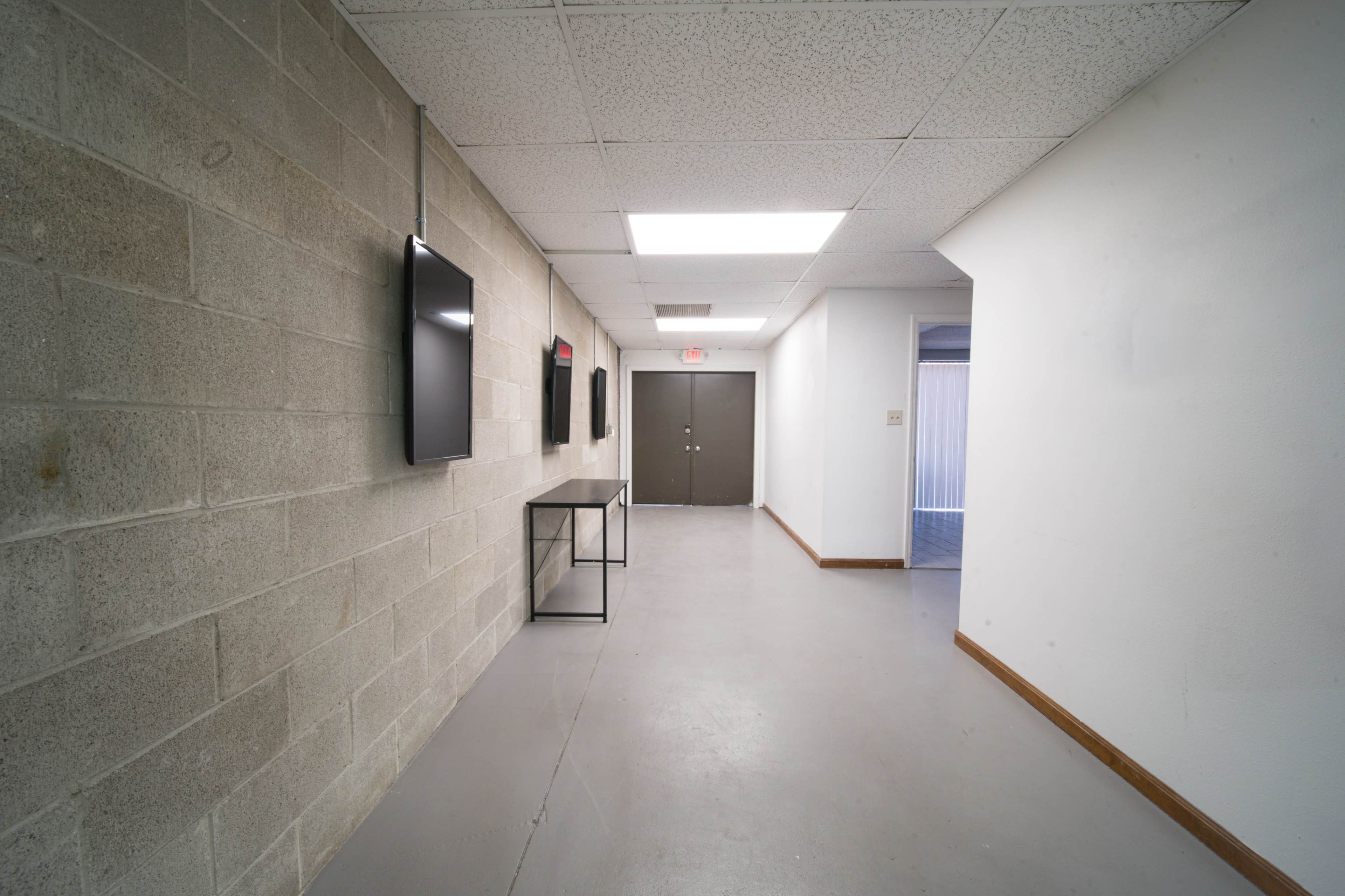 The image shows a long, narrow hallway with gray concrete walls, two wall-mounted televisions, and a door at the end.