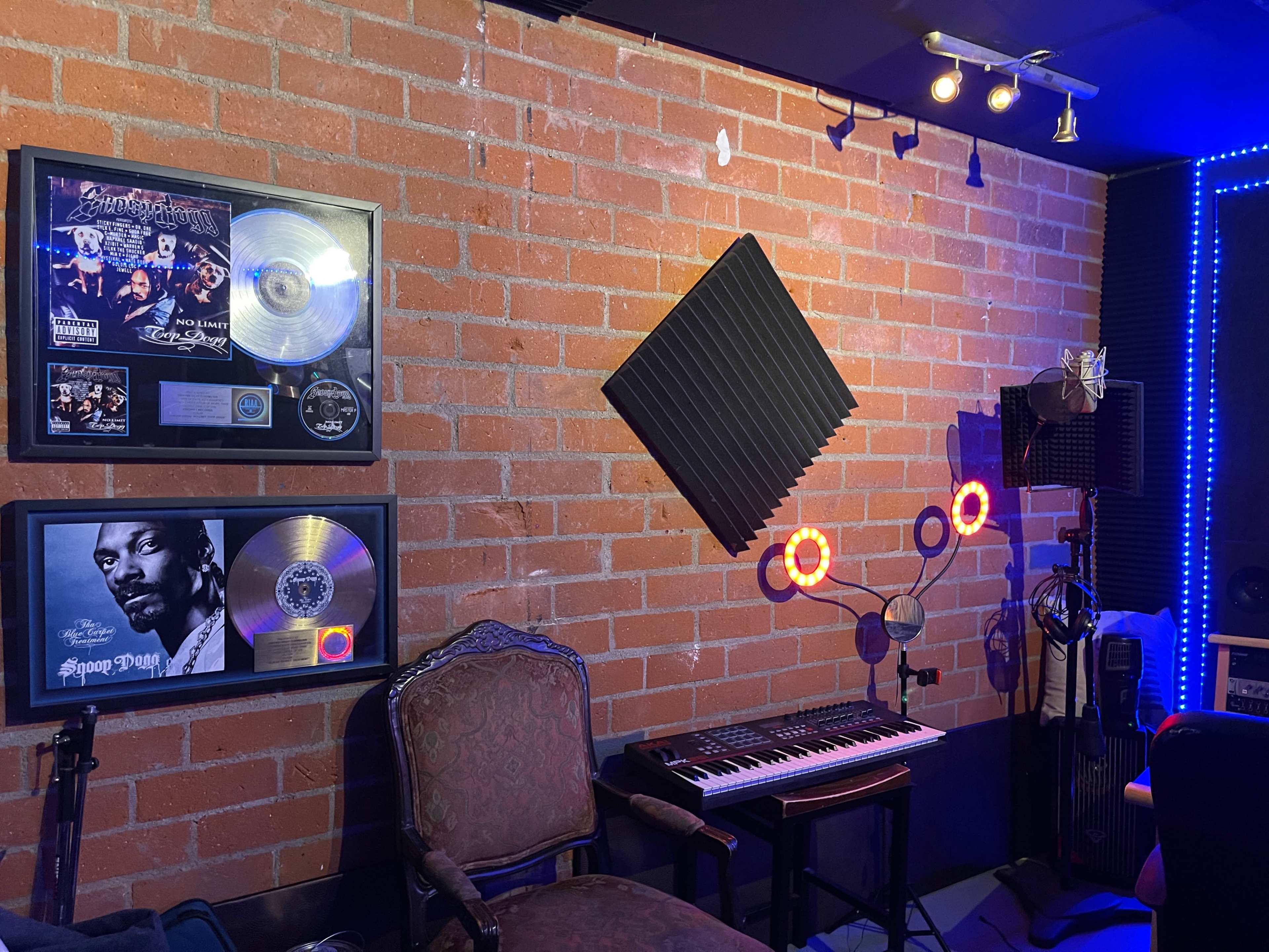 A music studio corner featuring framed platinum records on a brick wall, a keyboard, soundproof panels, and a microphone stand.