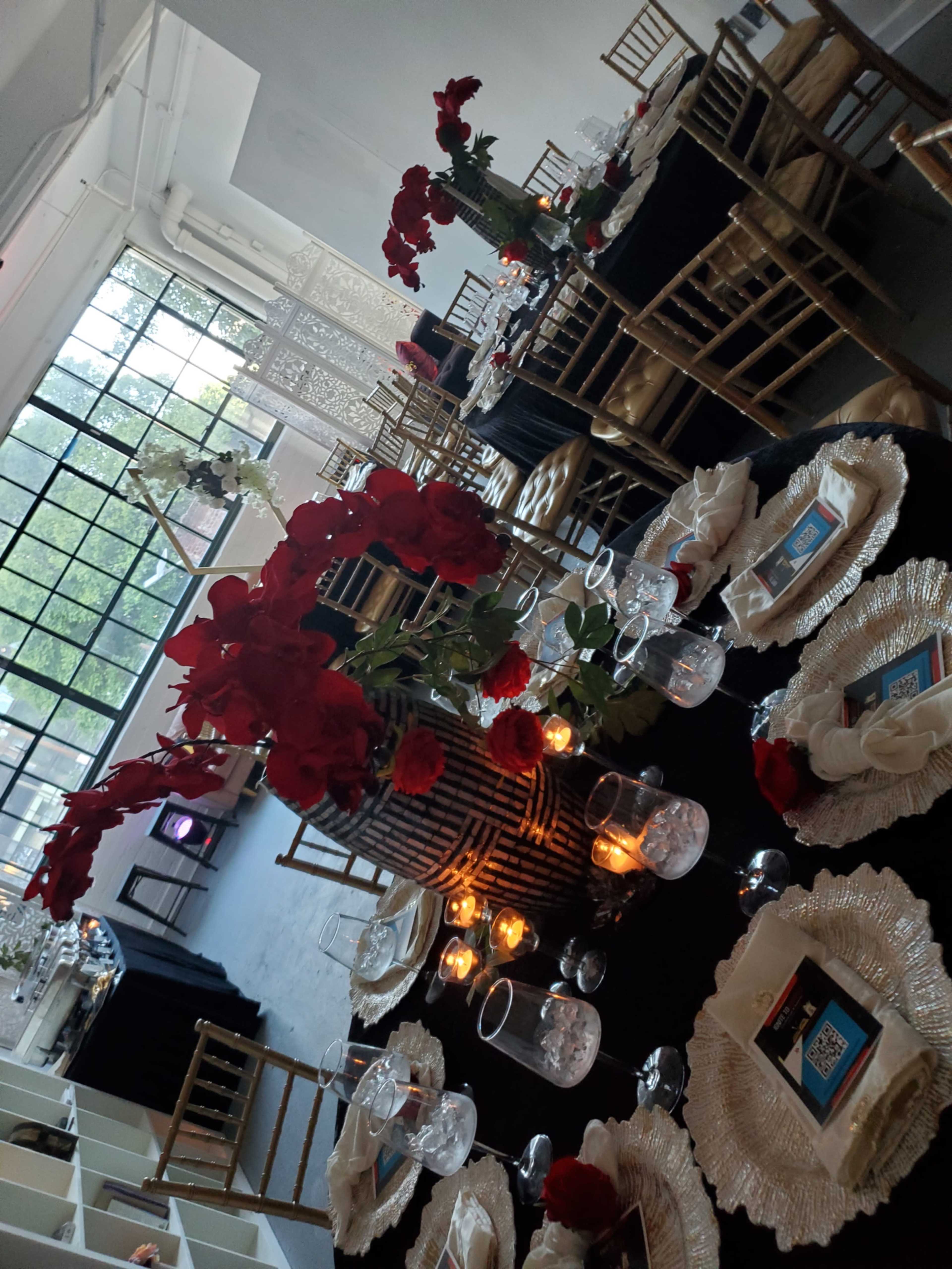 The image shows a elegantly arranged dining area with round tables covered in black and gold decorations, featuring floral centerpieces of red roses, candles, and place settings.