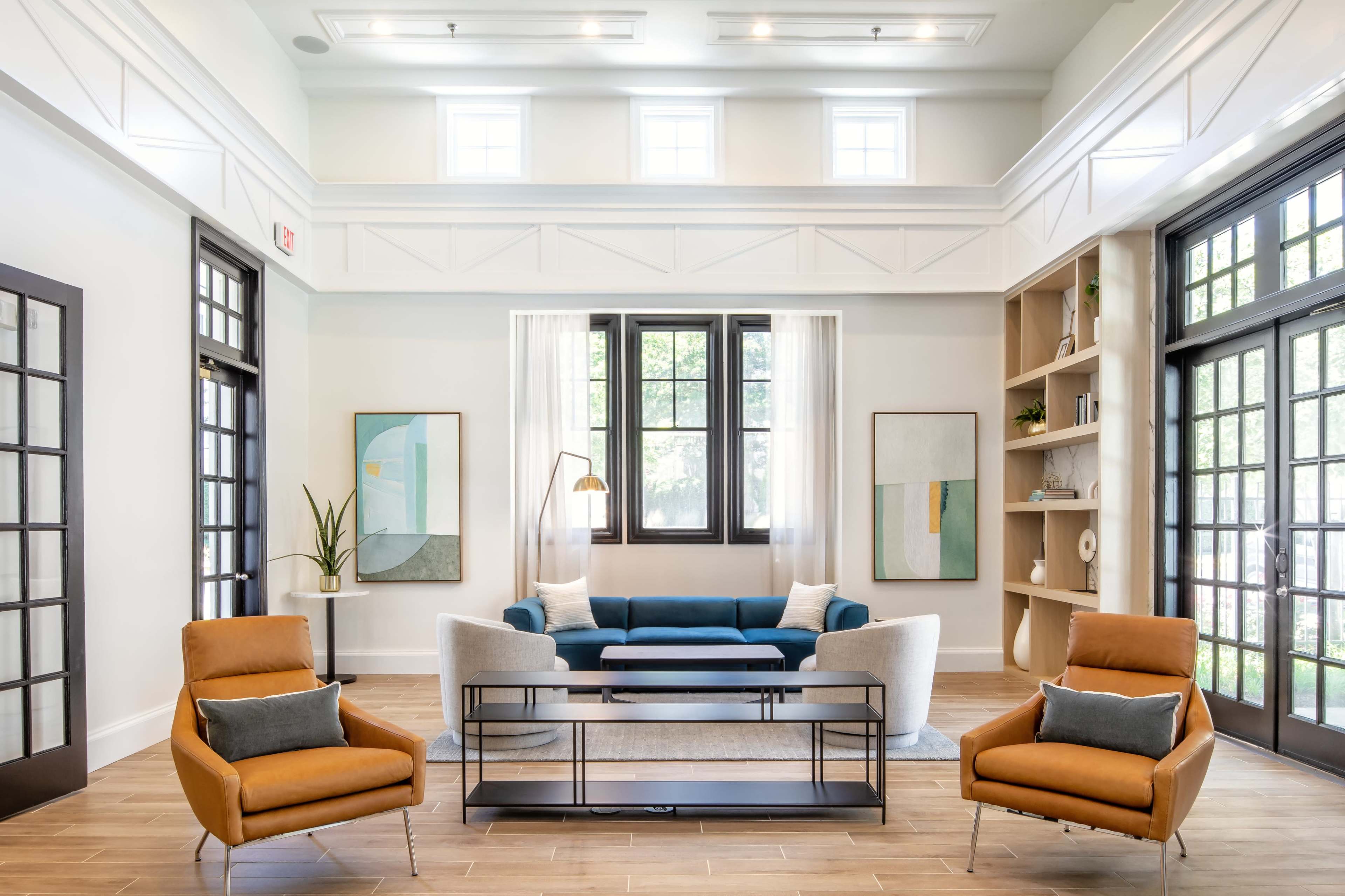 The image shows a modern living room with two orange chairs, a blue sofa, framed artwork on the walls, and large windows allowing natural light to enter.