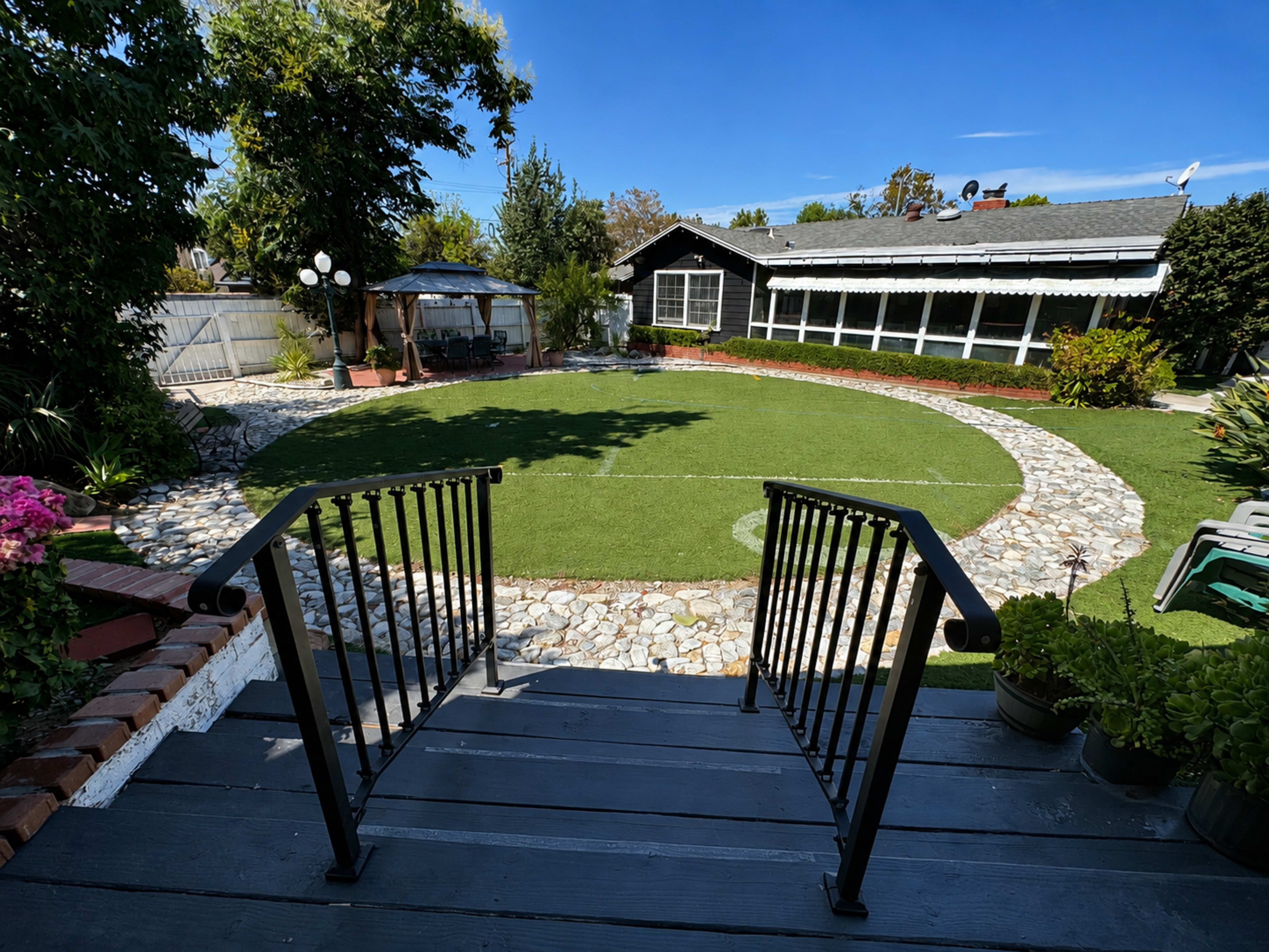 The image shows a landscaped backyard with a round patch of grass surrounded by stones, steps leading down from a deck, and a gazebo in the background.