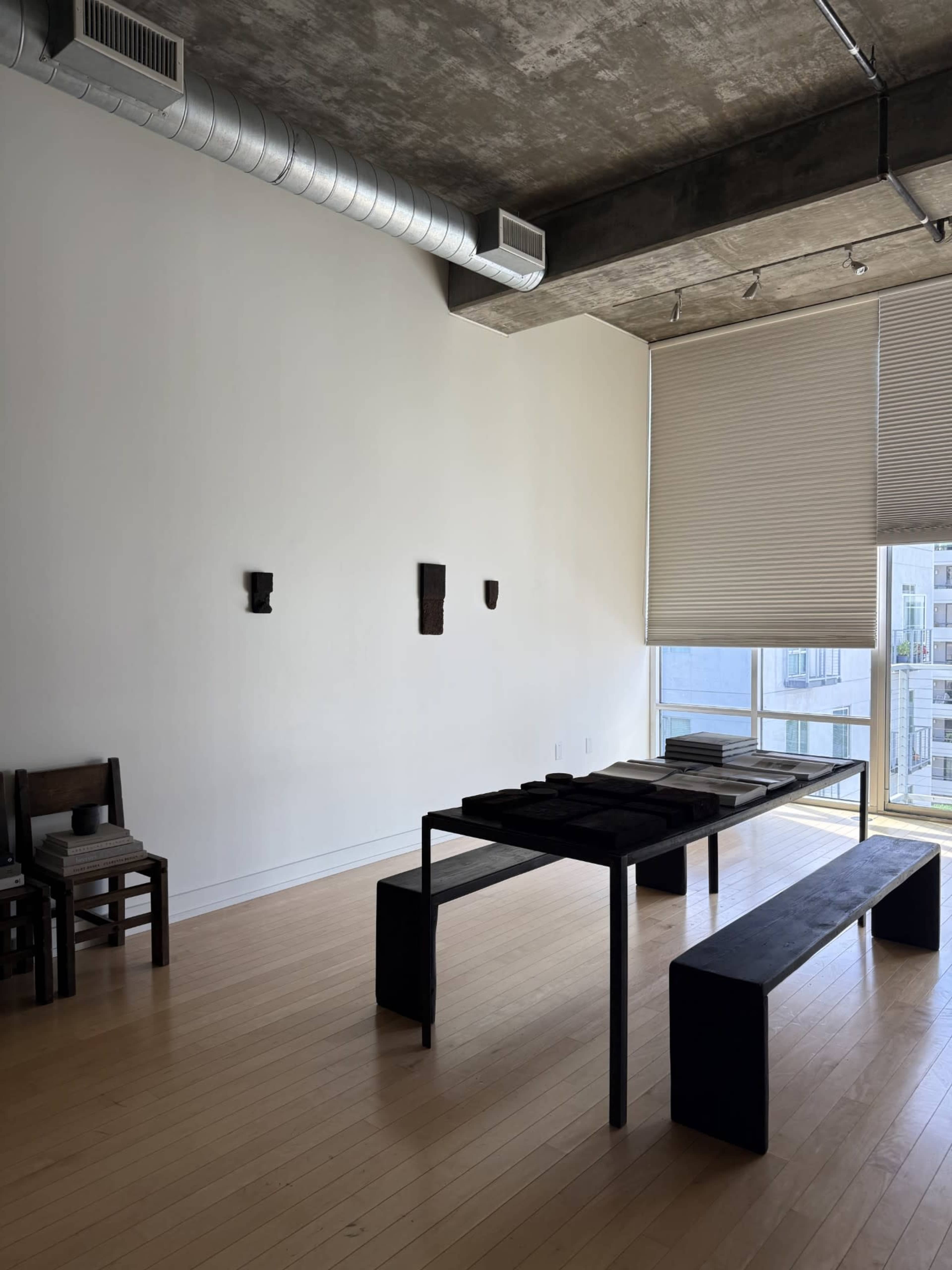 The image shows a minimalist interior space featuring a rectangular black table with several black items on it, surrounded by benches, and three small wall sculptures, with large windows allowing natural light to enter.