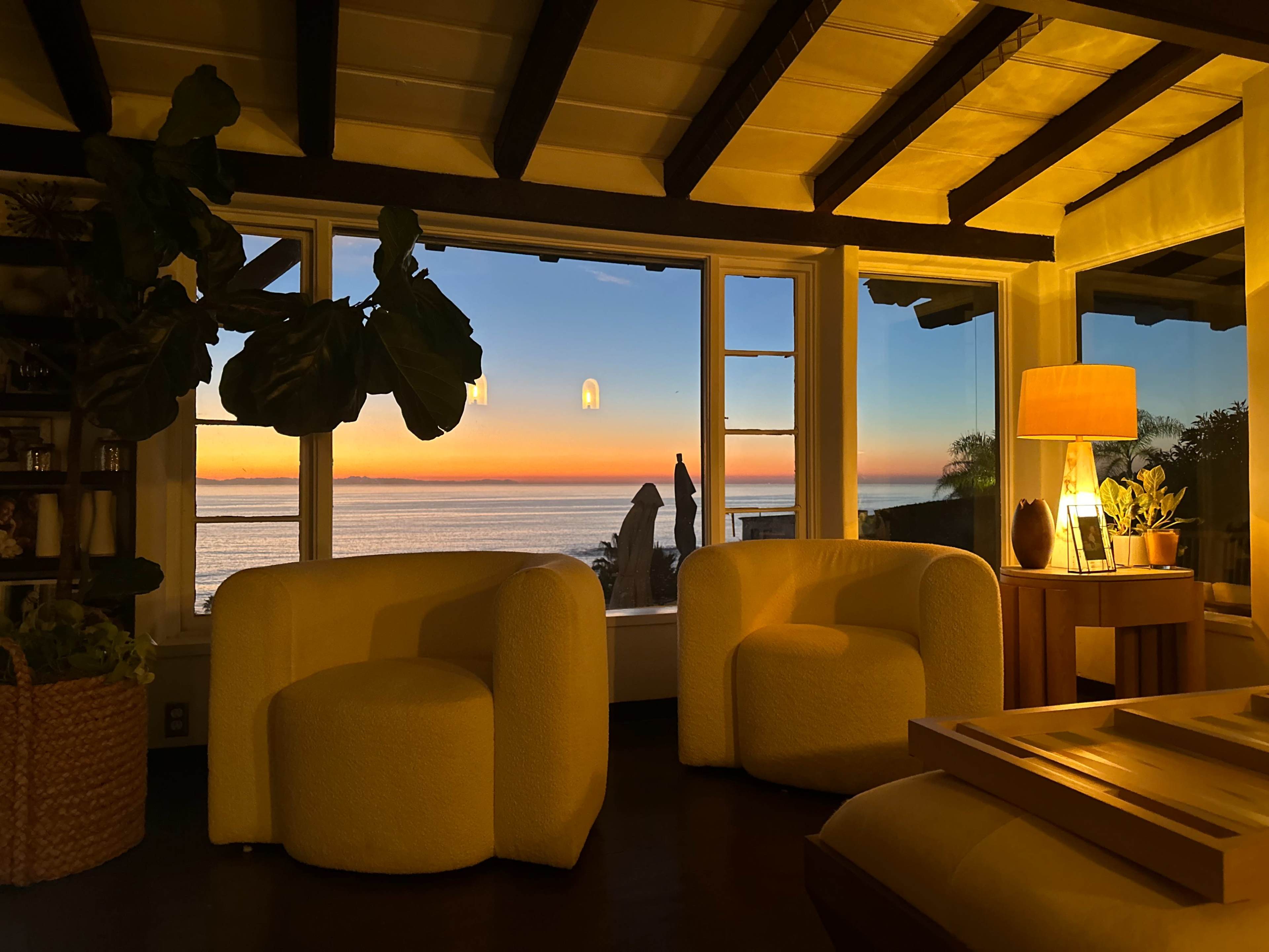 A cozy living room with two white chairs, a lamp on a wooden table, and large windows revealing a sunset over the ocean.