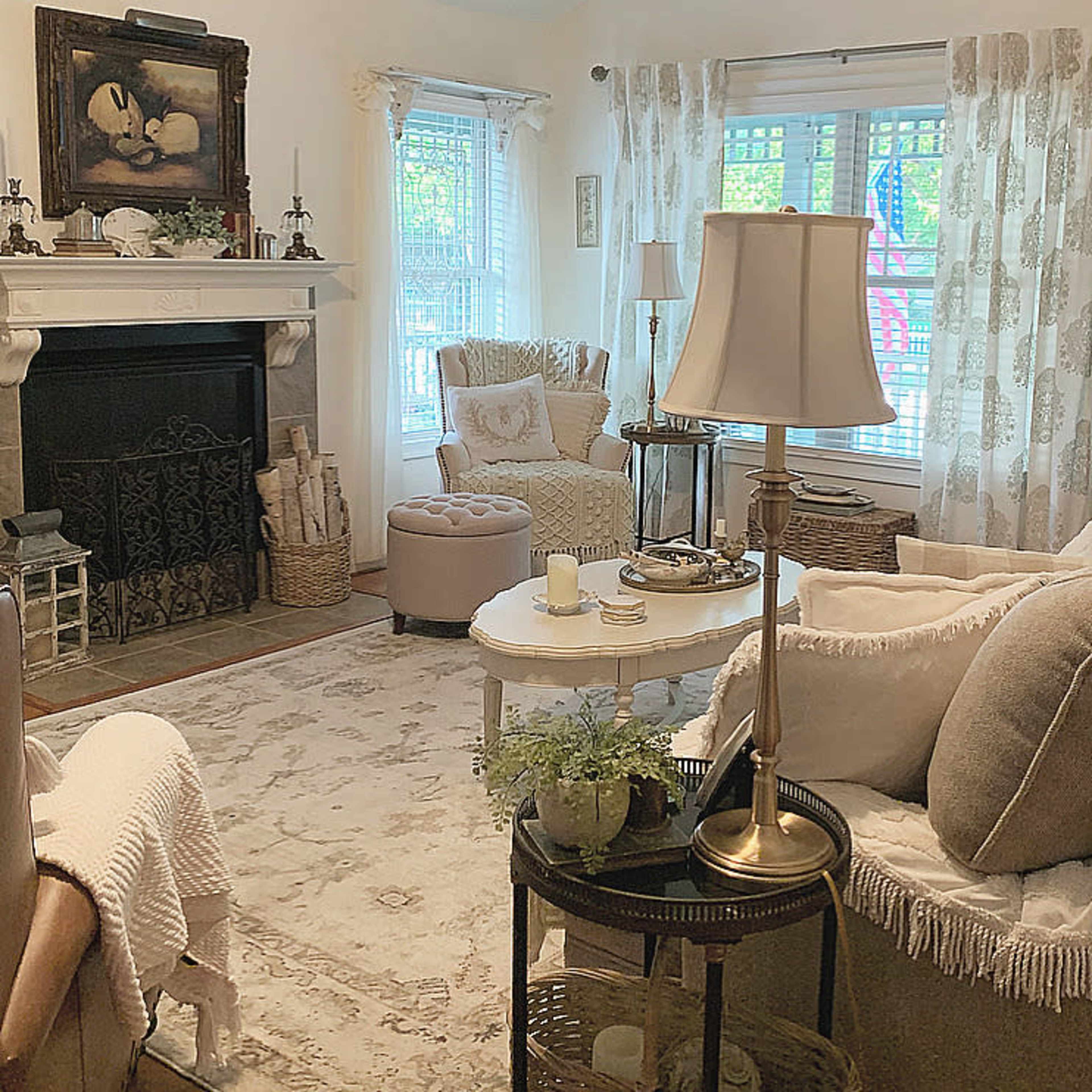 A cozy living room with a fireplace, a vintage chair, a round coffee table, and large windows dressed in sheer curtains.