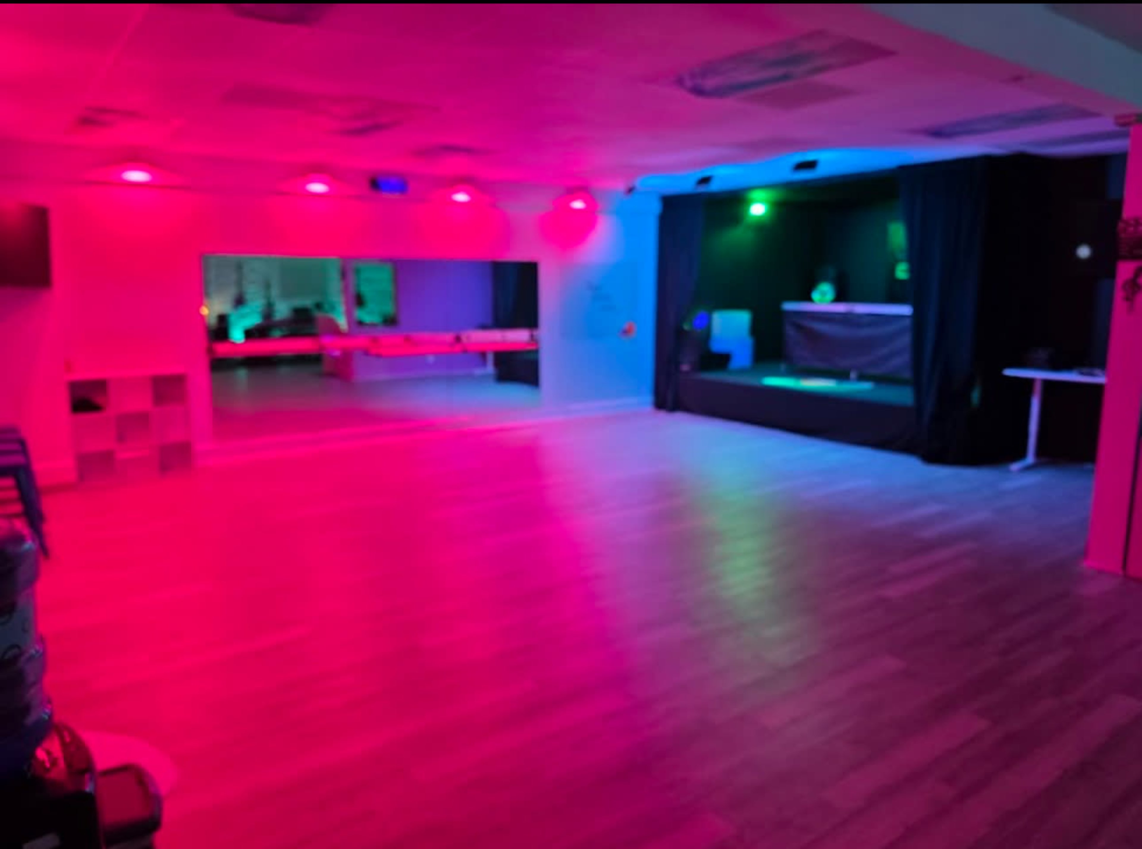 The image shows a spacious room with pink and green lighting, featuring a stage with a black curtain and a mirrored wall.