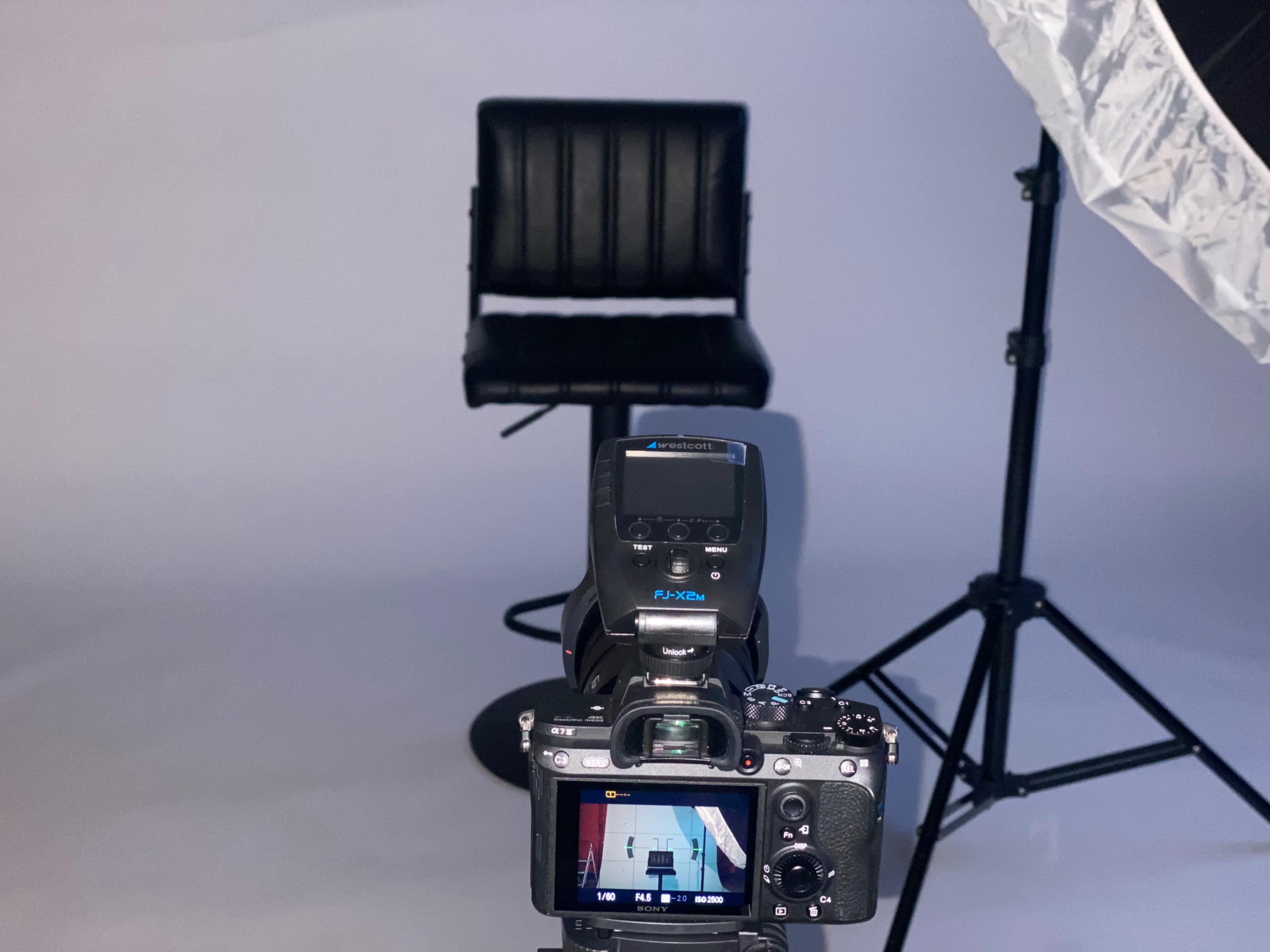 A camera is set up on a tripod facing a black leather stool against a plain backdrop, with a softbox lighting equipment nearby.