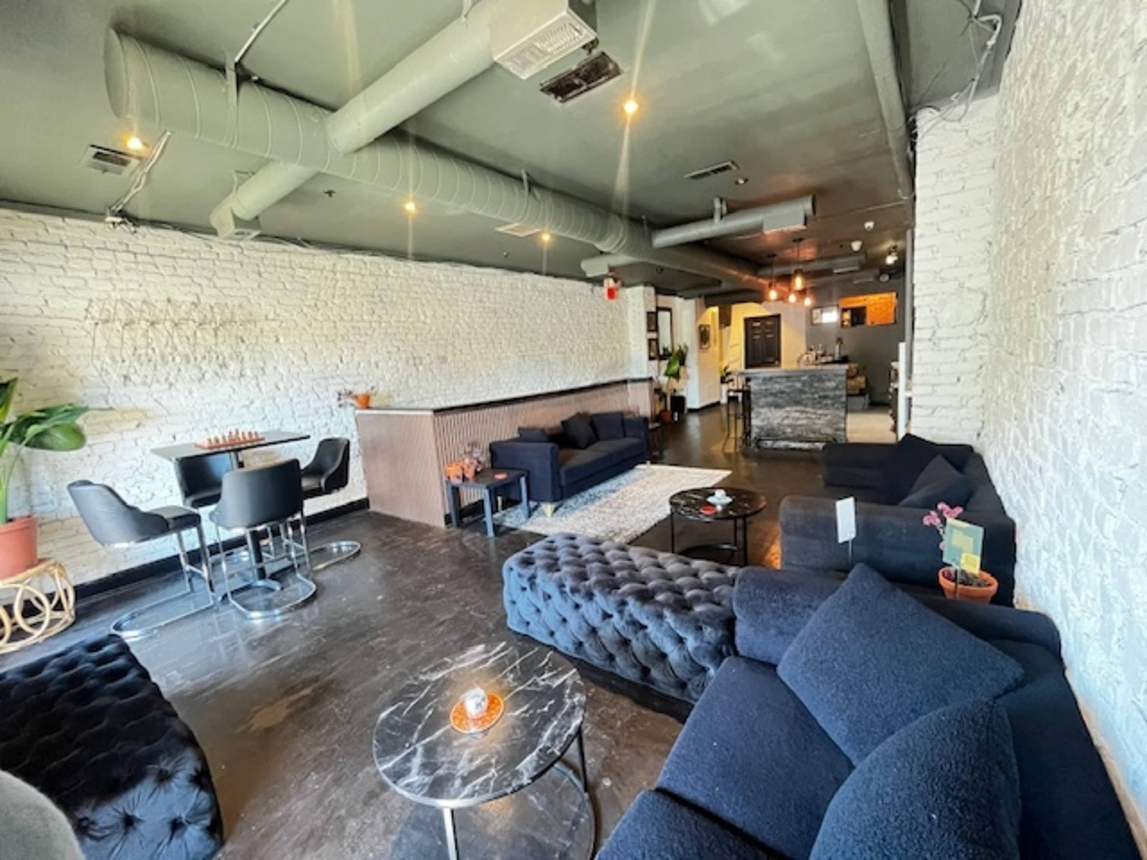 The image shows a modern lounge area featuring dark upholstered sofas, a black coffee table, and potted plants, with a restaurant counter visible in the background.
