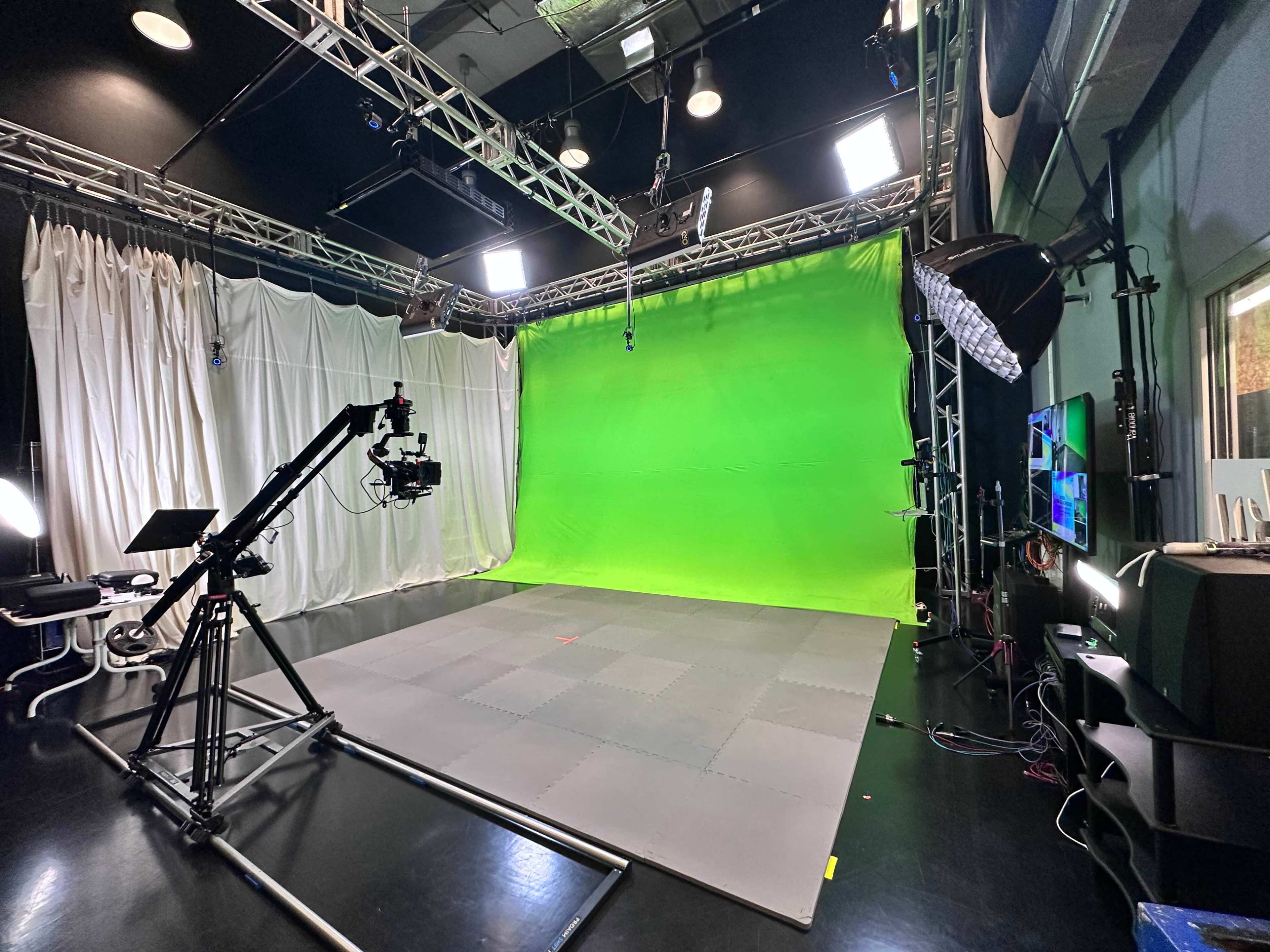 The image shows a film studio equipped with a green screen, lighting setup, and a camera on a tripod, situated on a gray tiled floor.
