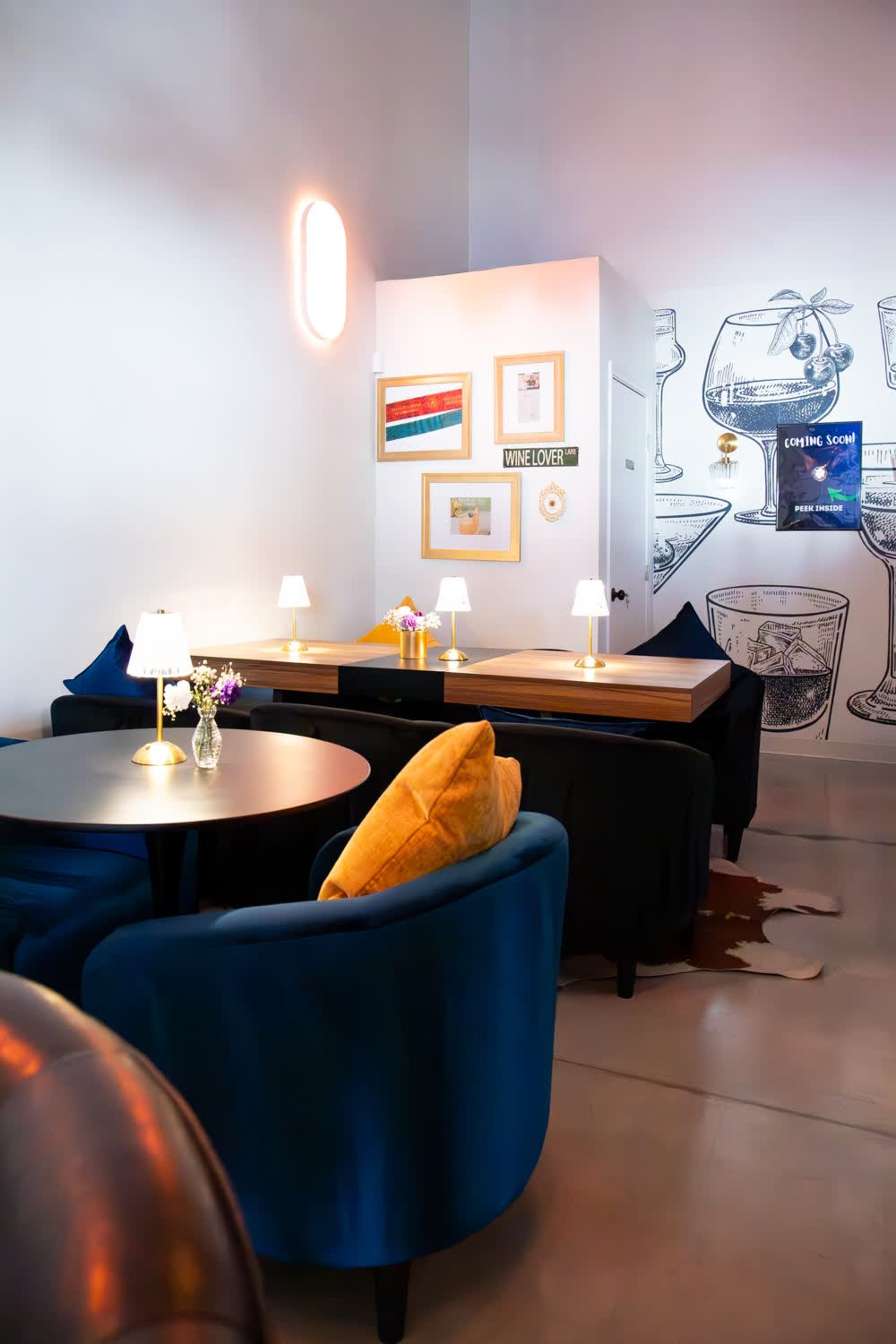 The image shows a modern café interior with blue velvet seating, small tables, and decorative wall art featuring various glassware.