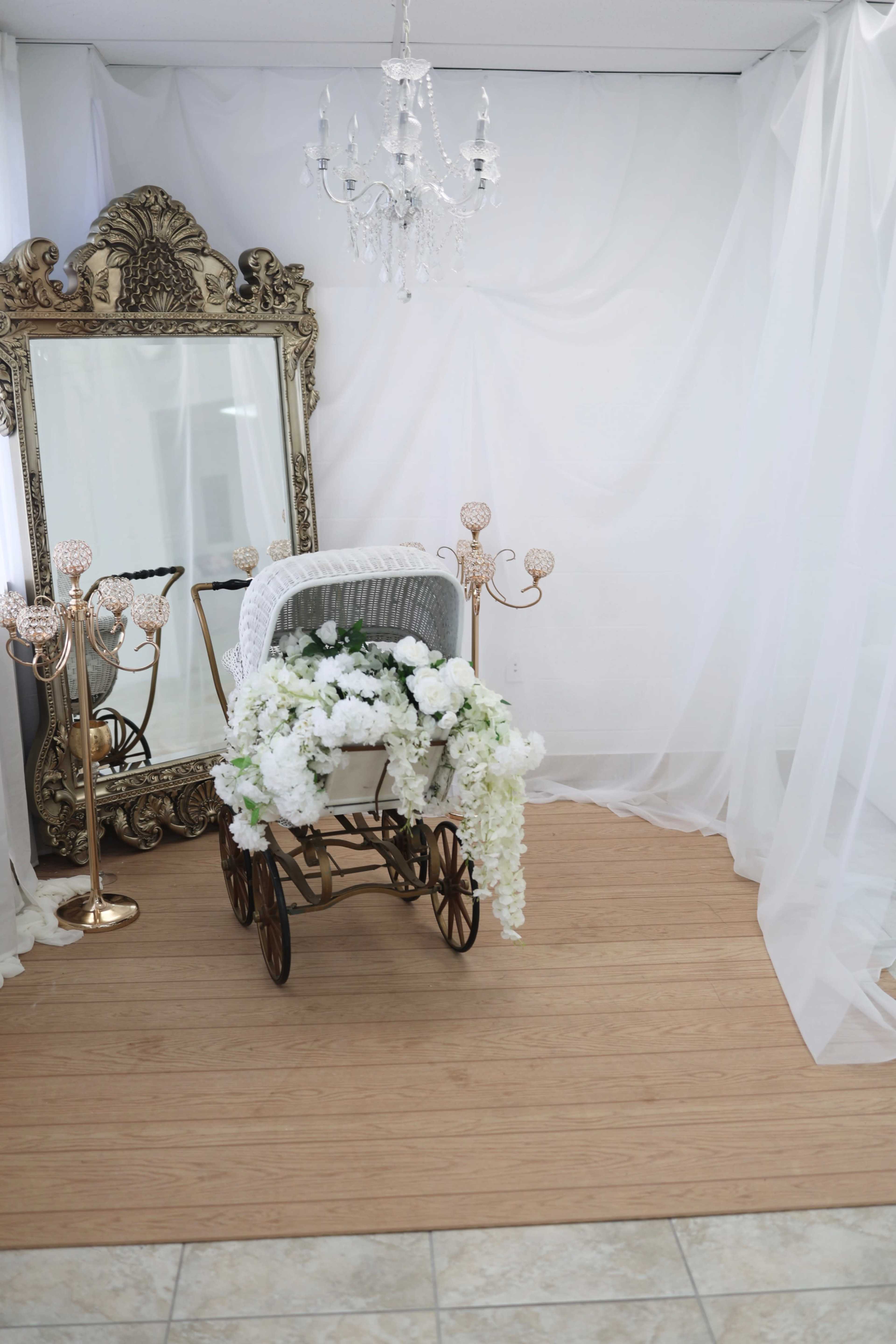 A vintage-style baby carriage adorned with white flowers is placed in a room featuring a large ornate mirror and a chandelier, all set against sheer white drapery.