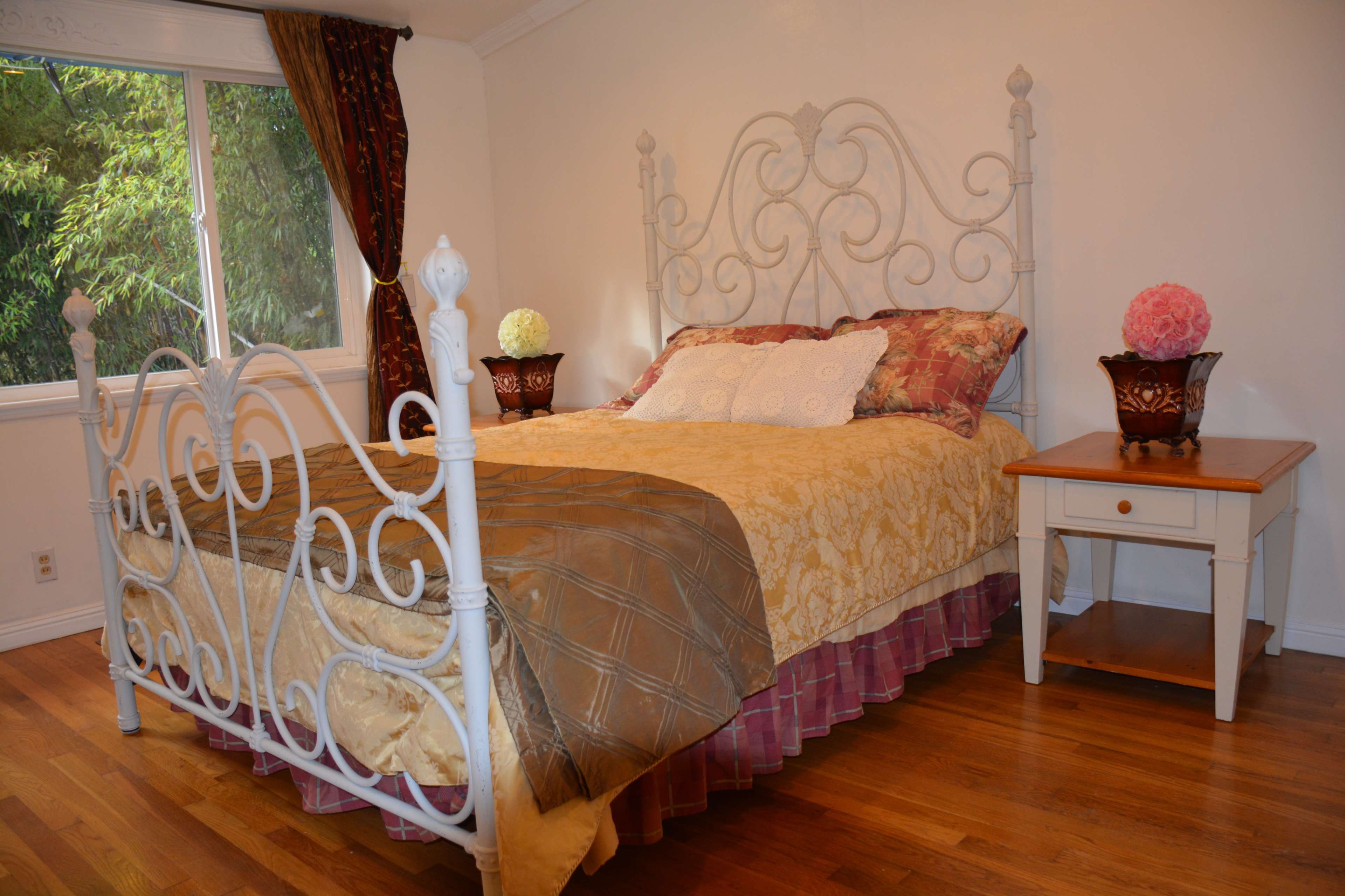 A bedroom featuring a metal bed frame with a patterned bedspread, a wooden nightstand, and large windows allowing natural light.