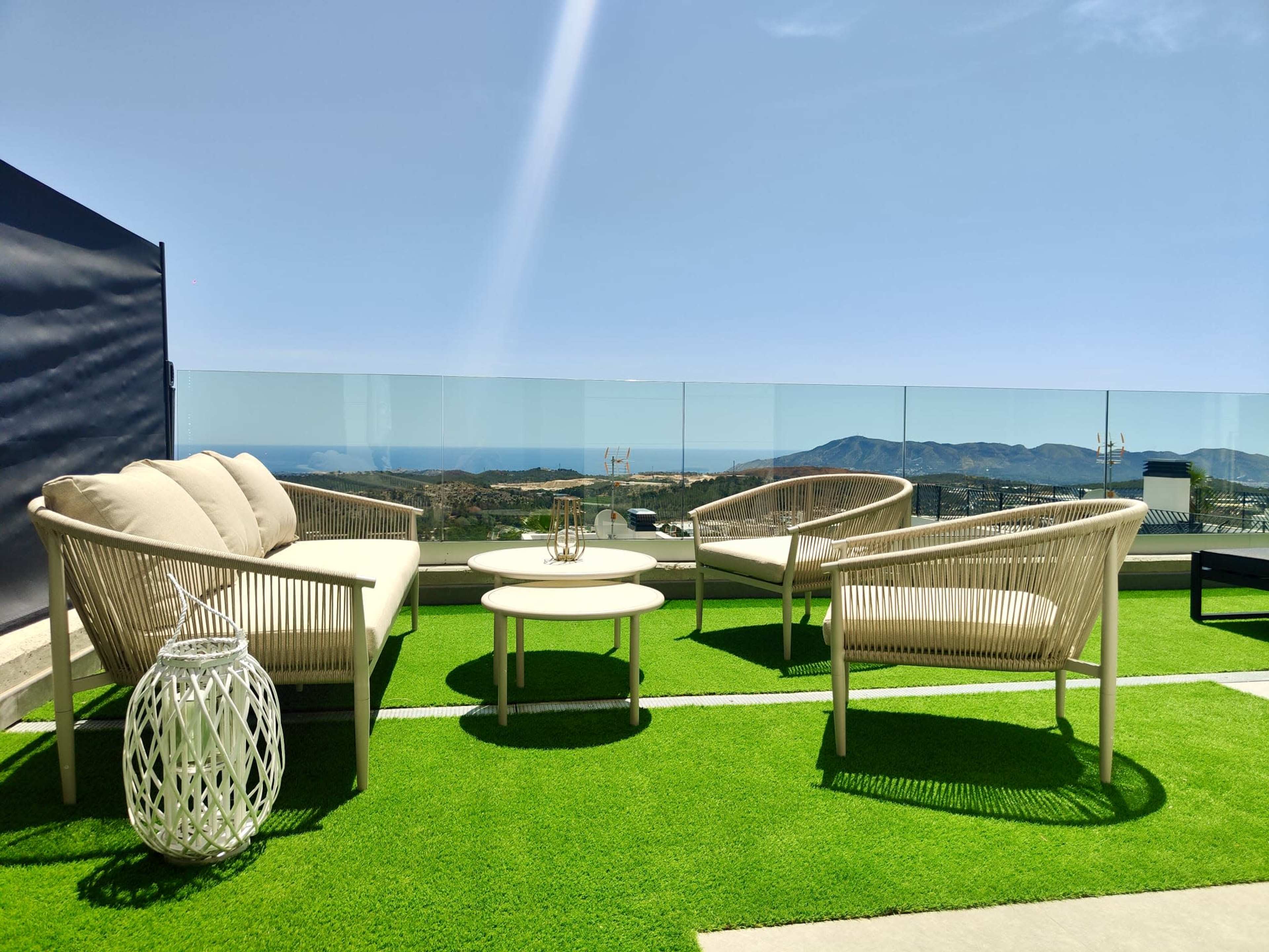 The image shows a rooftop terrace with two white chairs and a small round table on a green grass-like surface, overlooking distant mountains and the sea.