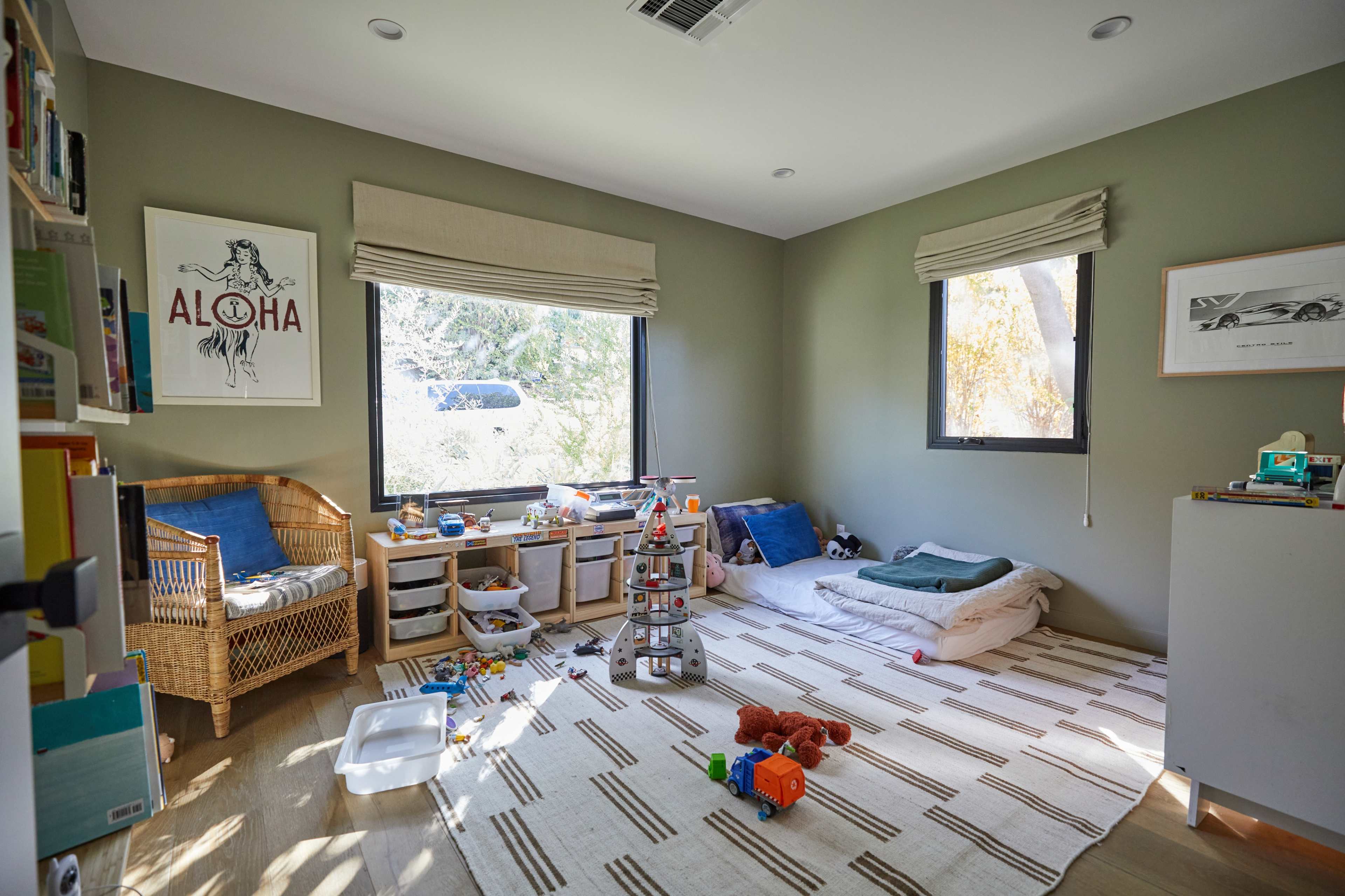 A playroom features a large rug, two windows with shades, a wicker chair, and various toys scattered on the floor.