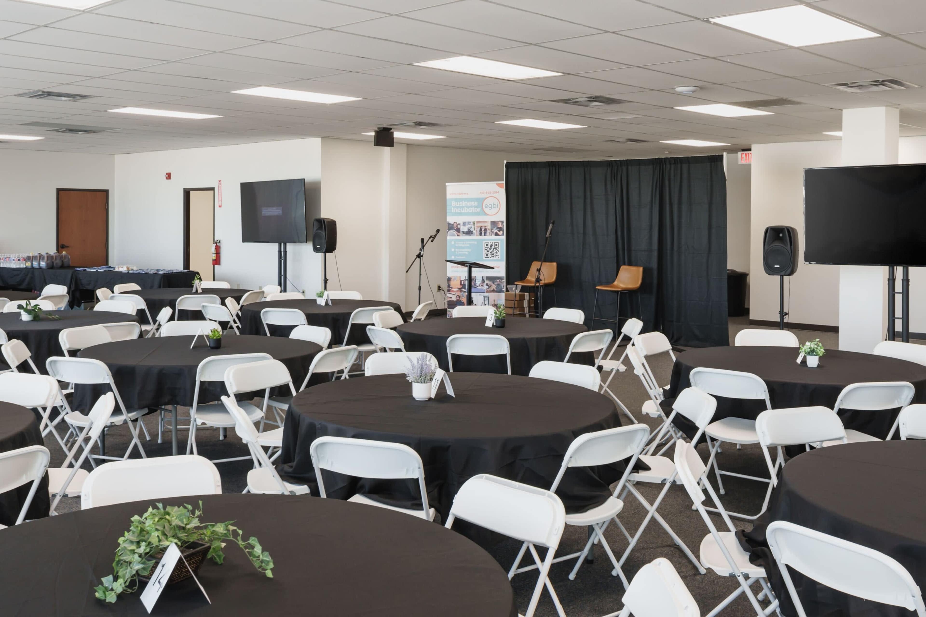 The image shows a large event space set up with several round tables covered in black tablecloths and arranged with chairs, while a stage area at the front features a microphone and two chairs.