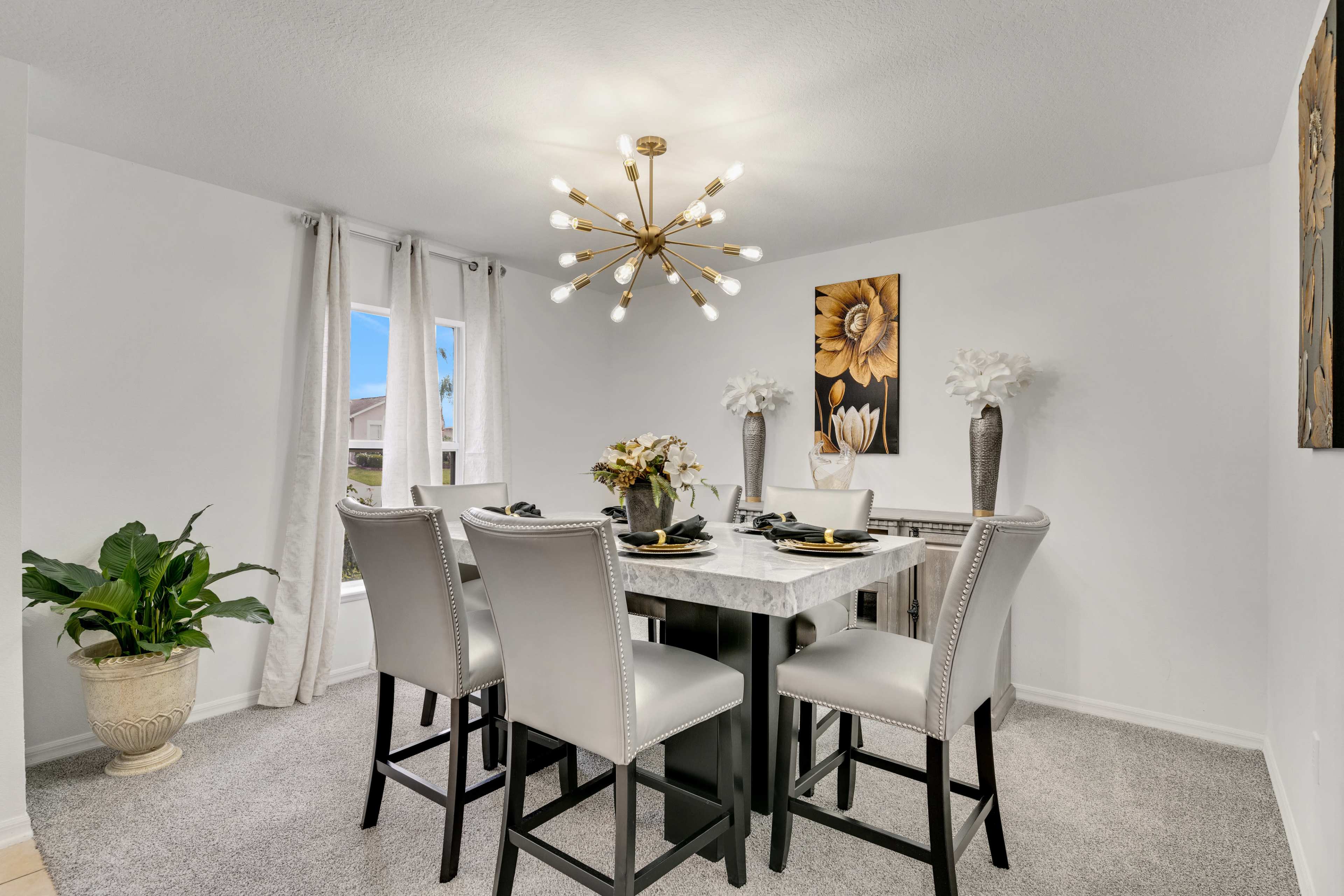 A modern dining area features a marble-top table surrounded by four upholstered chairs, illuminated by a chandelier, with decorative plants and artwork on the walls.