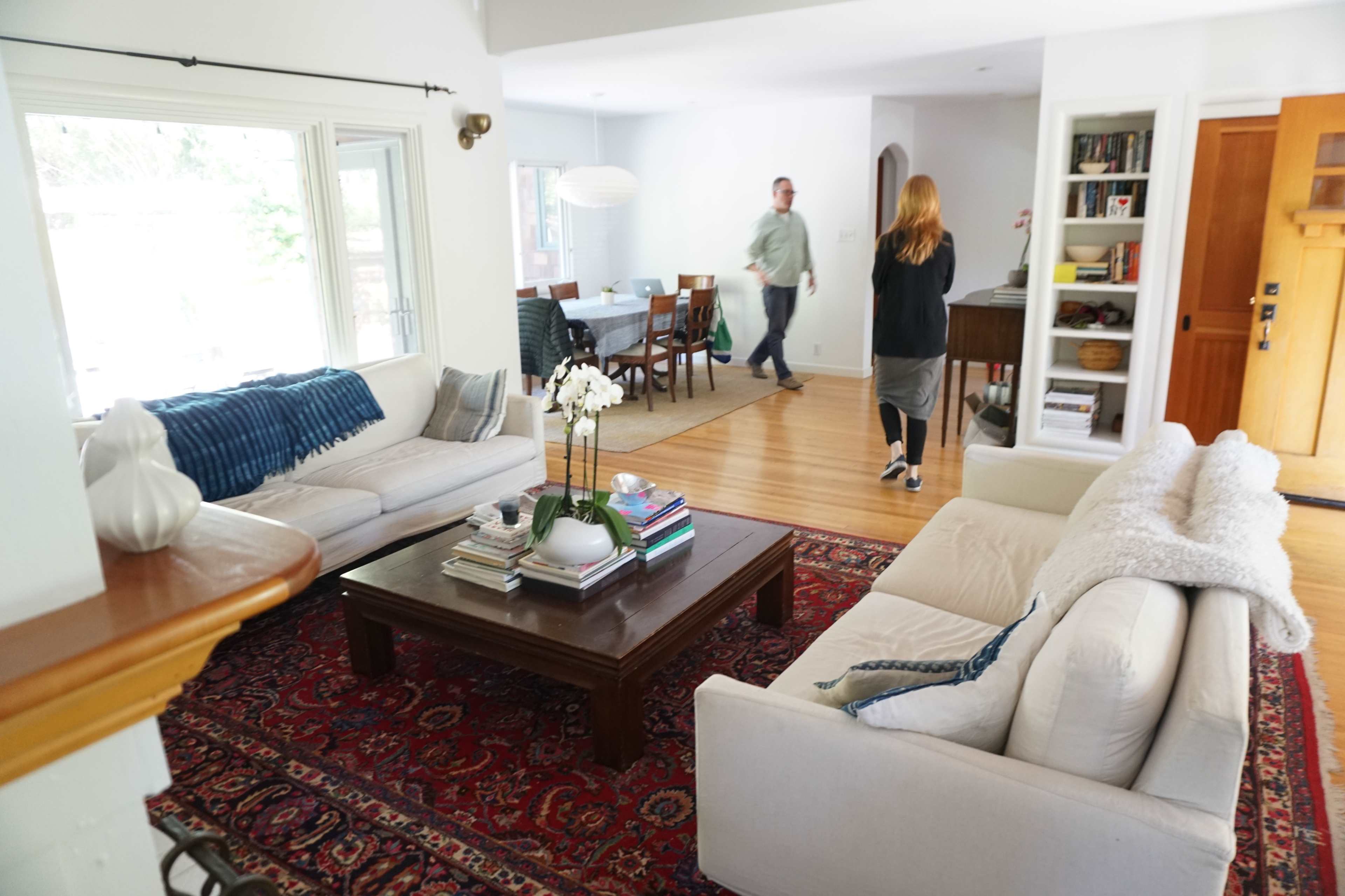 A living room with two sofas facing a coffee table, a bookshelf on one side, and a person walking towards the dining area in the background.