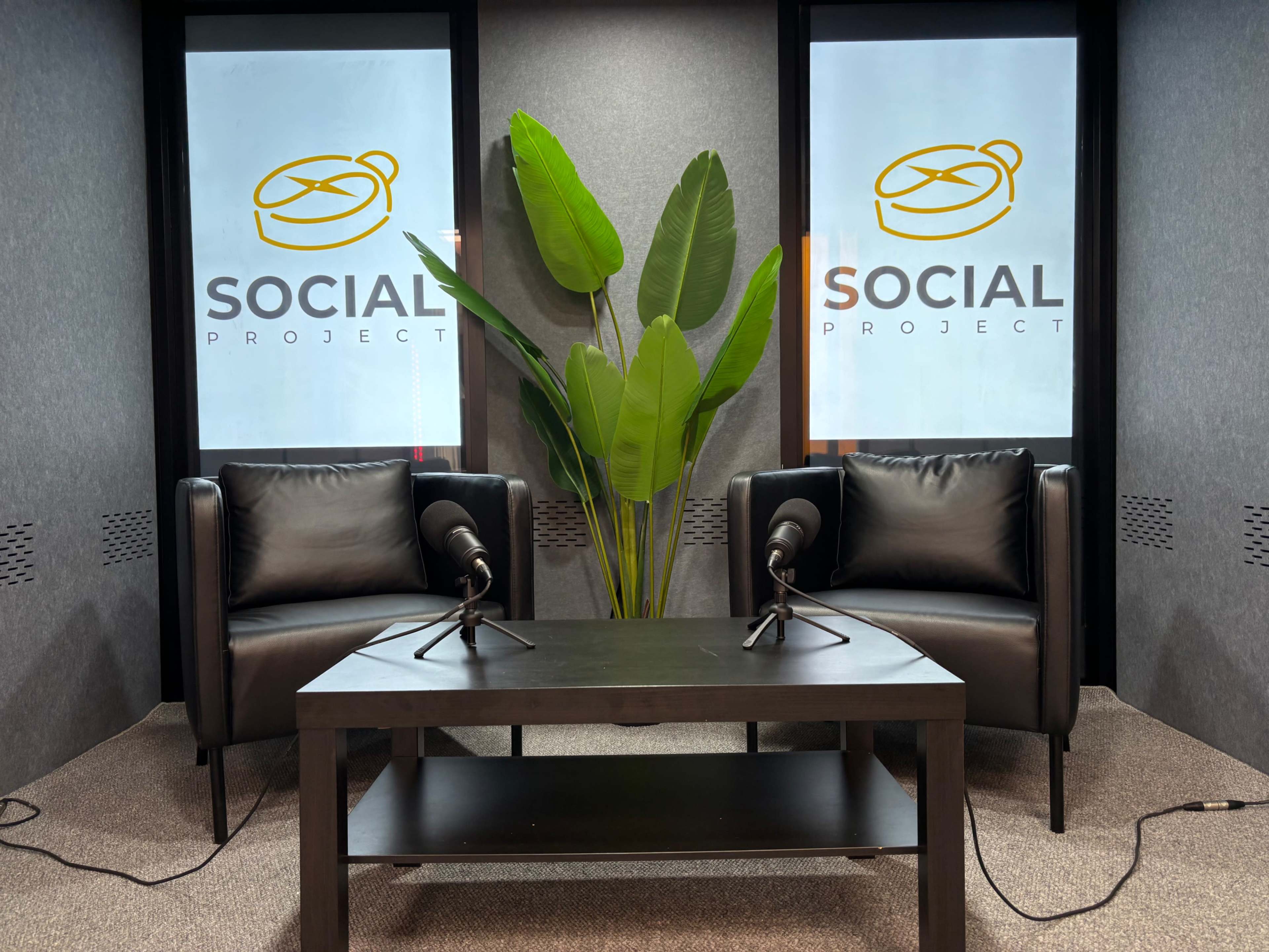 The image shows a modern studio setup with two black chairs, a small table, and large windows featuring the text "SOCIAL PROJECT" alongside a decorative plant.