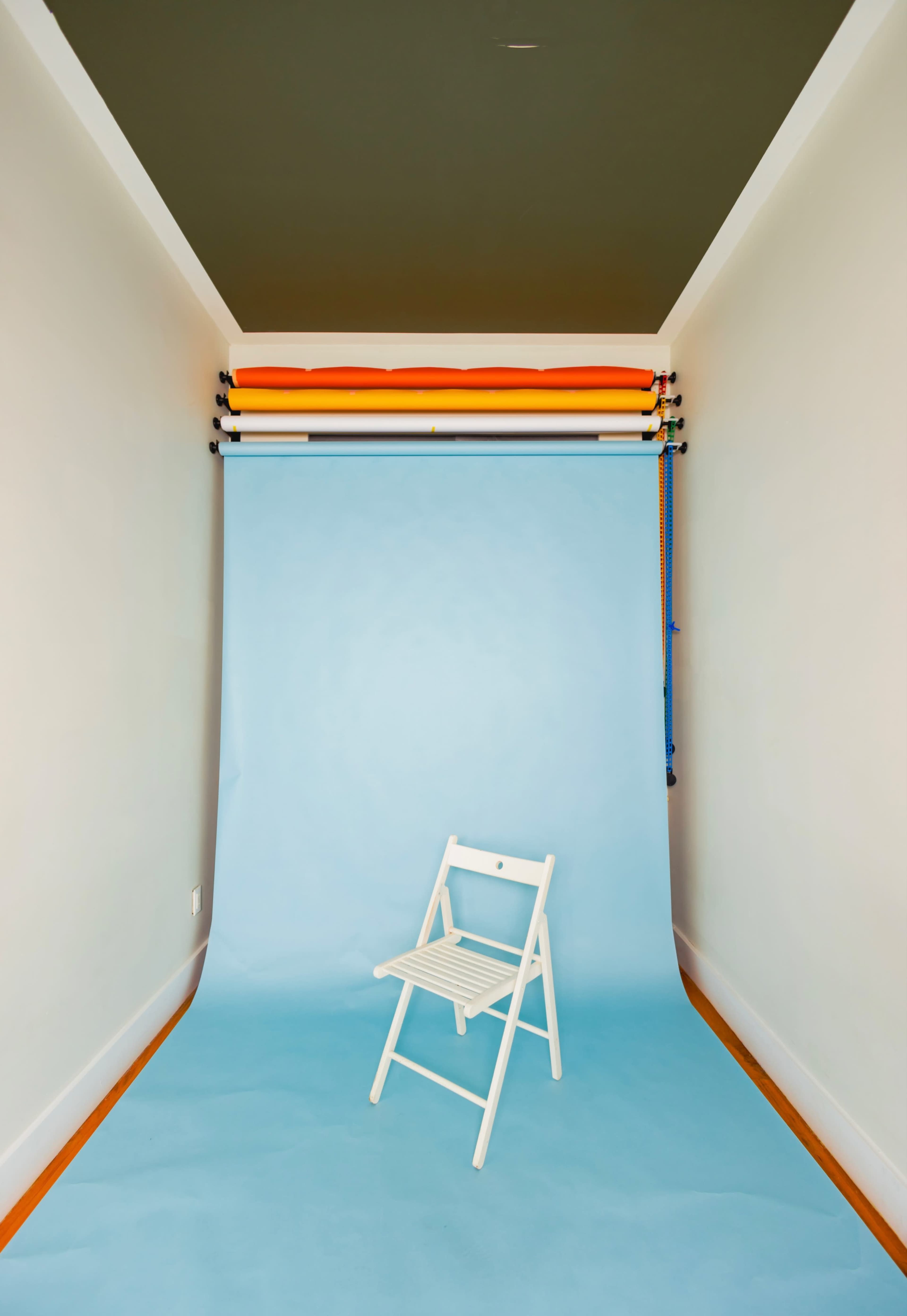 A minimalist studio setup with a light blue backdrop, a folded white chair in the foreground, and colored backdrop rolls mounted on the wall above.