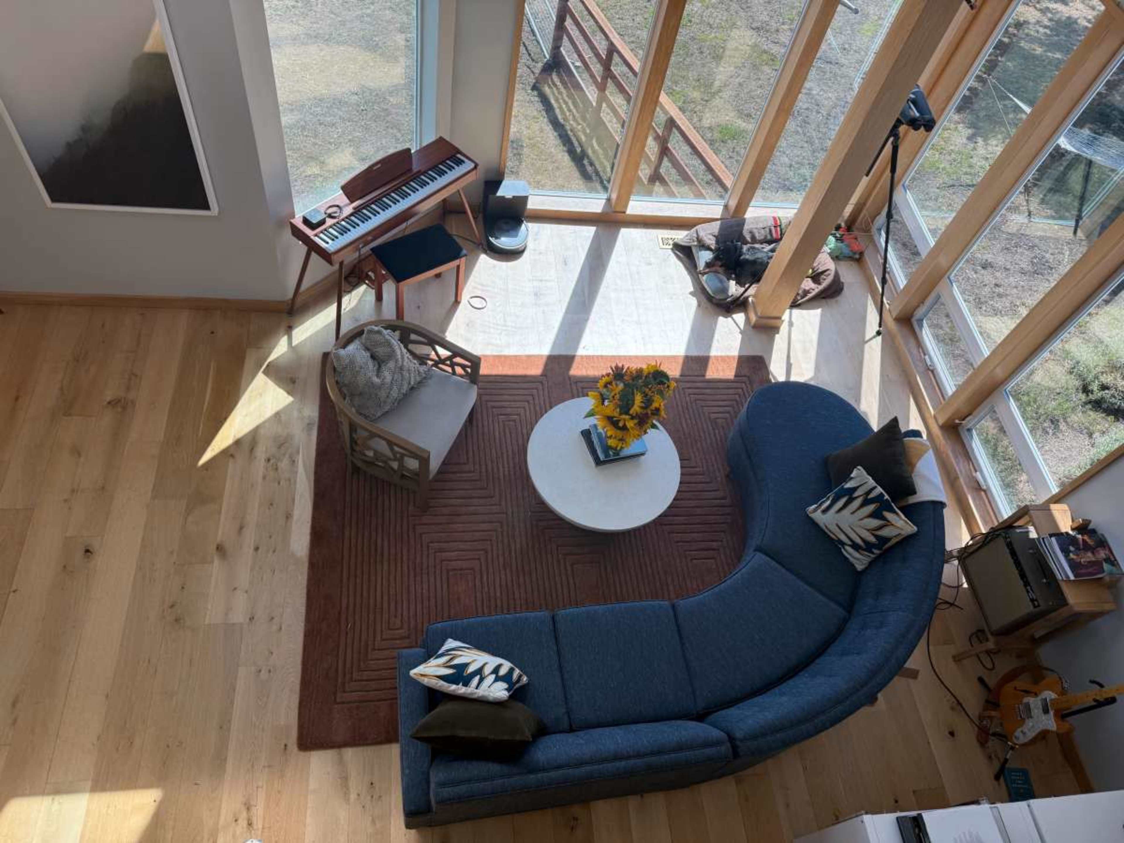 The image shows a modern, well-lit living room featuring a curved blue sofa, a round coffee table with flowers, and a keyboard against large windows.