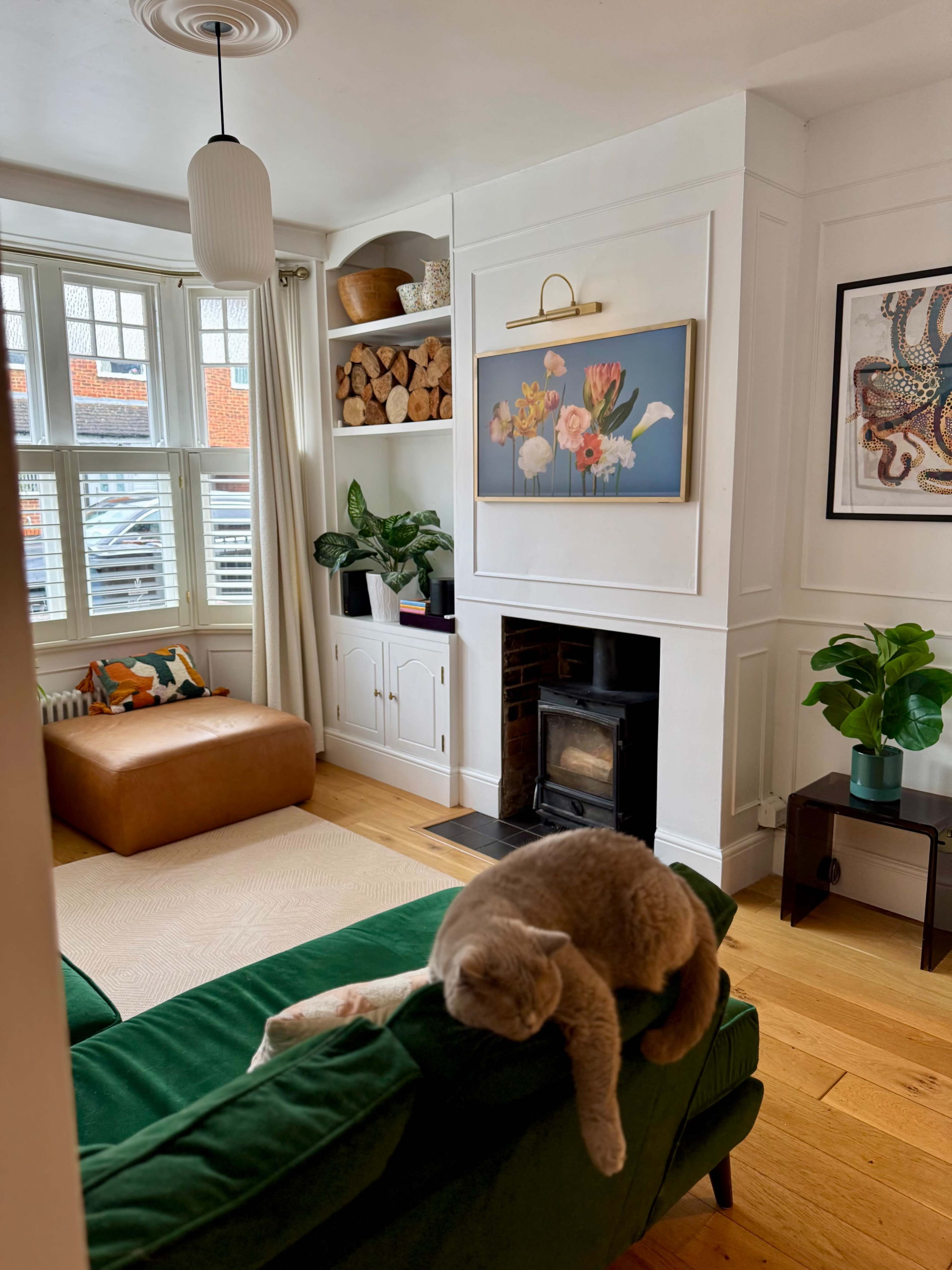 A gray cat rests on a green sofa in a cozy living room with a fireplace, a painting on the wall, and decorative shelves.