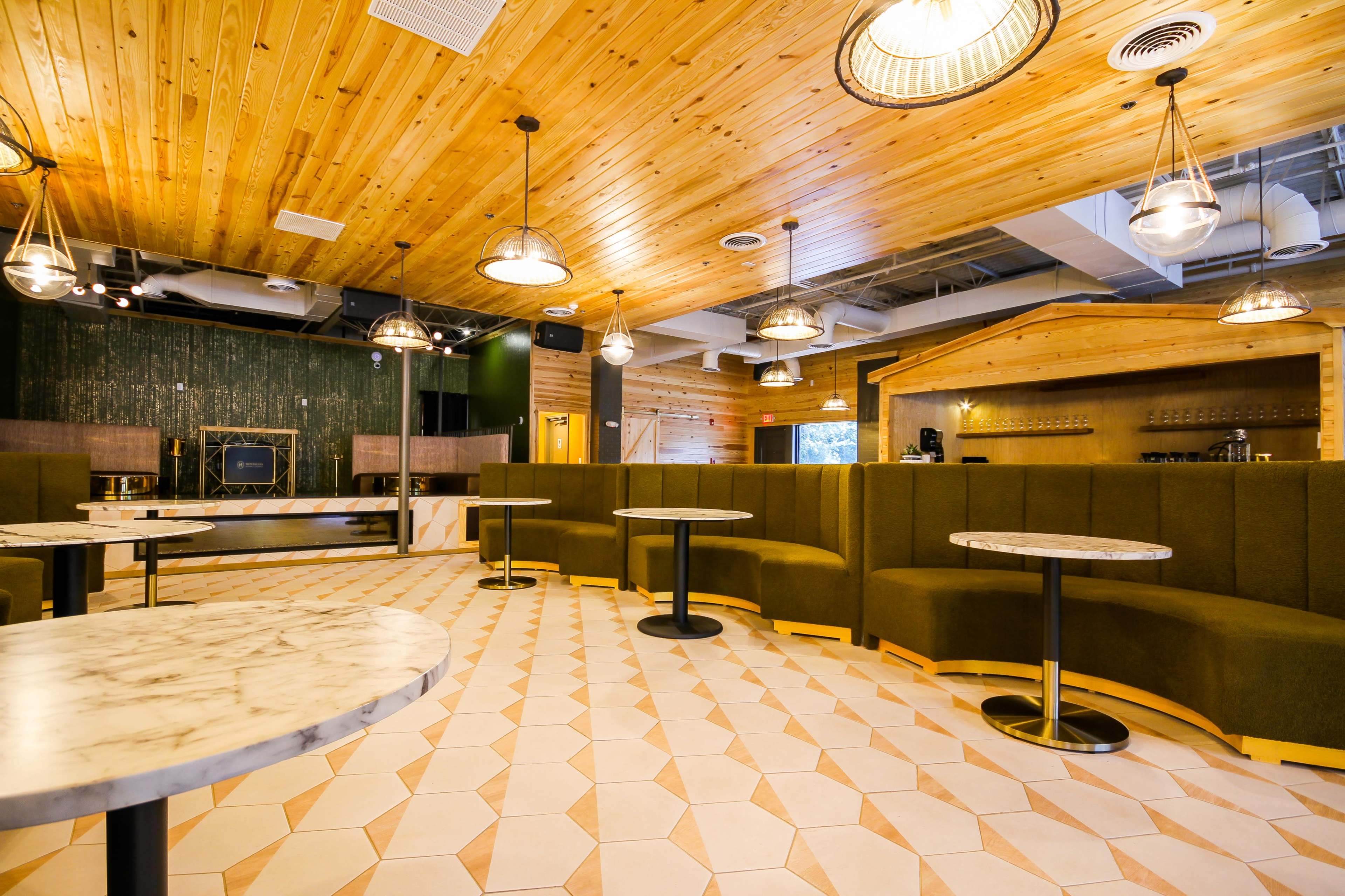 A modern café interior with wooden ceilings, green upholstered seating, and marble-topped tables arranged on a patterned tile floor.